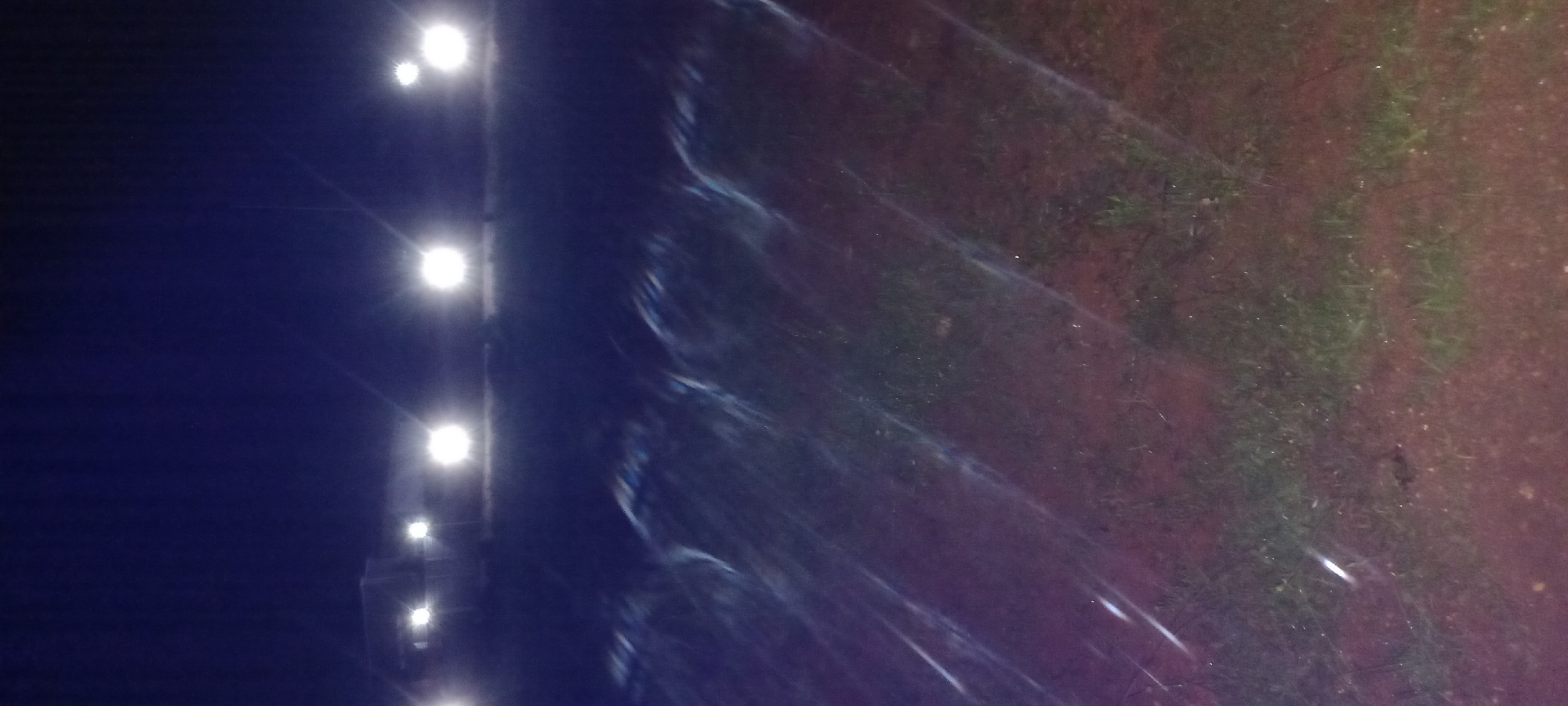 The image depicts a nighttime scene with strong artificial lighting. In the background, there are several bright, circular lights, likely from vehicles or a building. These lights cast strong reflections and glare onto the foreground, which appears to be a field or open area with some patches of grass and soil. The reflections create streaks of light across the image, indicating the presence of moisture or a reflective surface. The overall impression is that of an outdoor space illuminated by powerful lights at night. Without further information or context, the specific location in Kwang, Nigeria cannot be determined from the image alone.