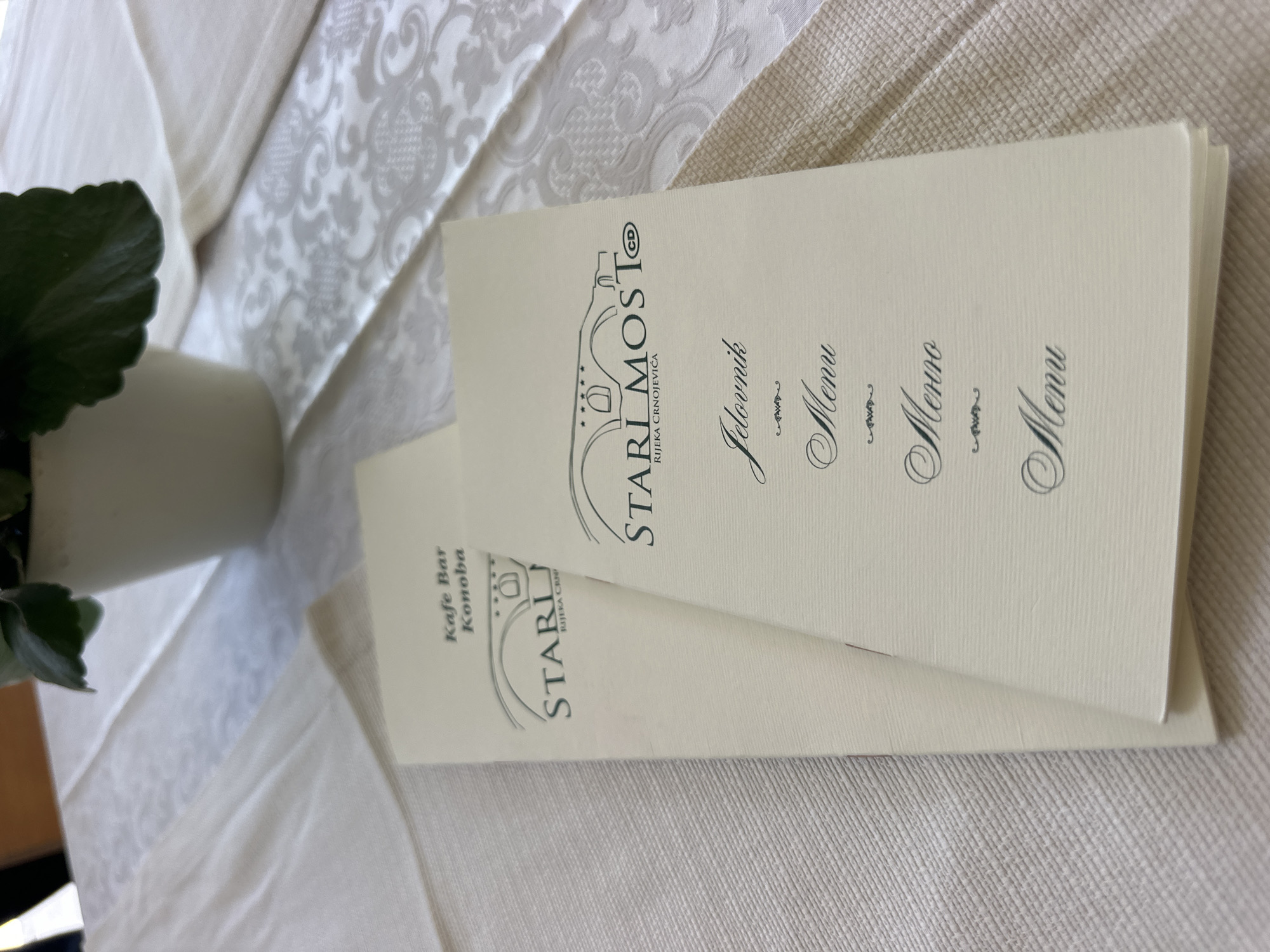 The image shows two menus on a table set with a white, textured tablecloth. One menu is partially obscured by the other. Both menus are a light cream color and feature elegant, dark script. The top menu displays a logo with a bridge and the text "STARI MOST@" and "RIJEKA CRNOJEVIĆA". Below the logo, the word "Jelovnik" is prominently featured, followed by "Menu" in a slightly smaller font. The second menu, peeking out from underneath, also has a similar logo with "STARI MOST" and "RIJEKA CRNOJEVICA" and the text "Kafe Bar Konoba". The menus are angled on the table, and a green leaf from a plant is visible in the lower left corner, suggesting a restaurant or cafe setting. The lighting appears to be natural, indicating it might be daytime. The overall impression is one of a well-presented dining establishment in Rijeka Crnojevića.