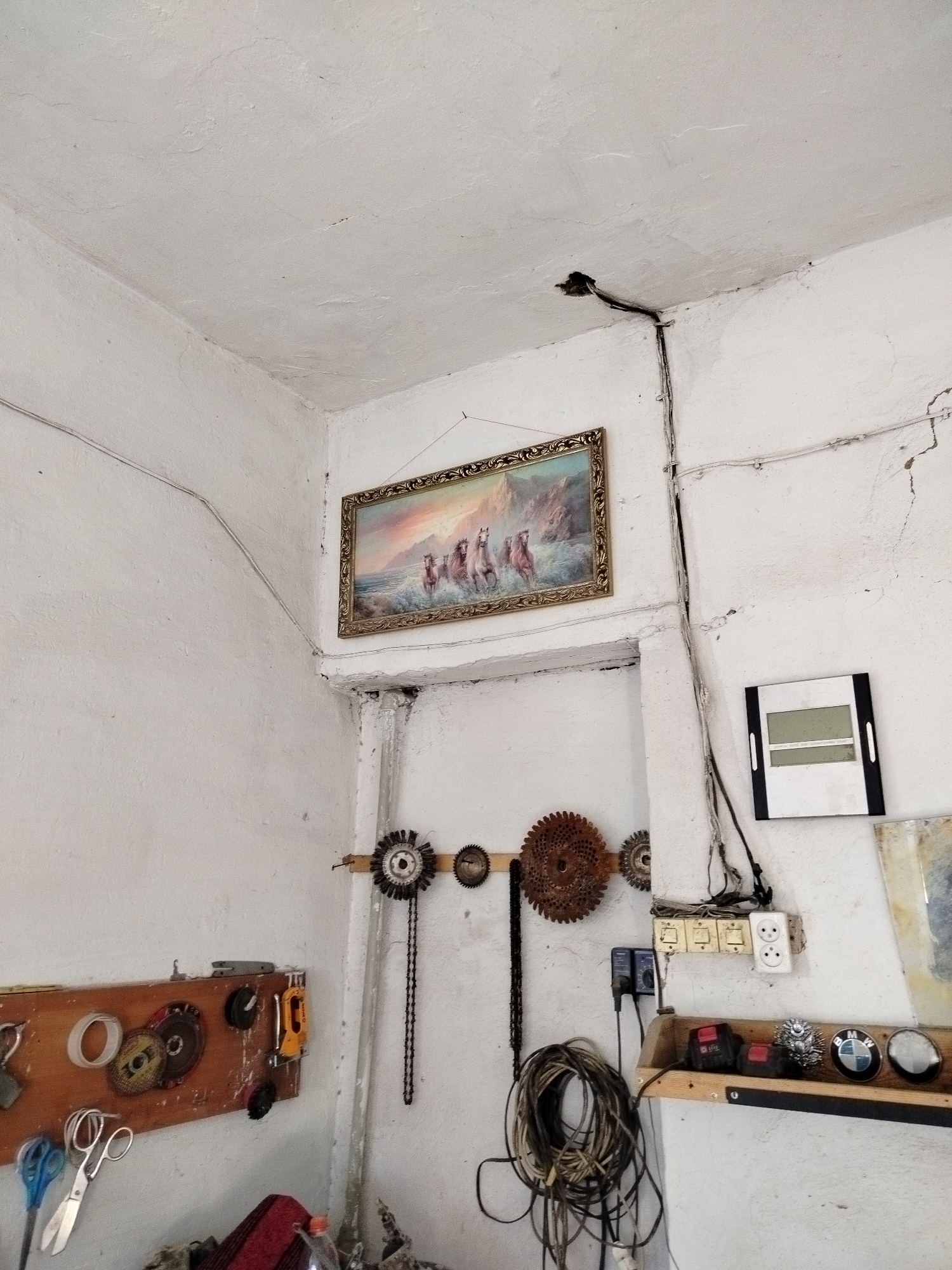 The image depicts a cluttered interior corner of a room, possibly a workshop or garage, in Kölkent, Kazakhstan. The walls are painted white and show signs of wear, including cracks and staining, particularly near the ceiling. A framed painting of horses running on a beach hangs on the wall in the upper left quadrant. Electrical wires are visible running along the ceiling and down the wall, connected to several switches and outlets.
Various tools and workshop items are organized on shelves and wall-mounted boards. On the left, a wooden board displays rolls of tape, cutting discs, and a stapler. Below this, a pair of scissors, one blue and one silver, lies on a surface. To the right of the painting, a circular wire brush attached to a chain hangs on the wall, along with other gears and circular metal objects.
Further to the right, a shelf holds electrical components, including what appear to be power adapters and coiled electrical wires. Another shelf below this contains various items, including what seem to be car emblems, one of which is identifiable as a BMW logo. The overall impression is of a functional but unkempt workspace.