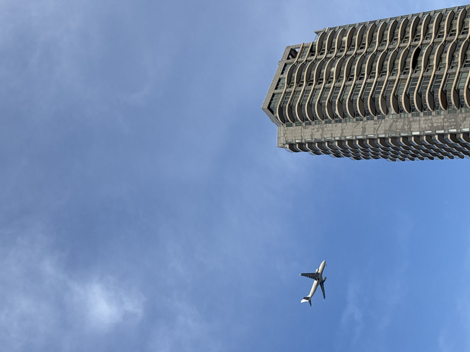 The image displays a clear daytime scene in Tel-Aviv, Israel, featuring a tall modern residential building and a commercial aircraft against a bright blue sky.
The prominent structure on the right side of the frame is a high-rise building, consistent with the architecture of the Neve Tzedek Tower or similar adjacent high-rises in Tel-Aviv. It is constructed from grey-toned materials, characterized by numerous floors with distinctive, curvilinear, light-colored balconies that undulate along the visible facade. Dark windows are visible behind the balconies.
In the mid-left portion of the sky, a white passenger jet is observed in horizontal flight, moving from left to right across the frame. The aircraft has its landing gear extended, suggesting it is either on approach for landing or has recently taken off, likely from Ben Gurion Airport (TLV). Its underside appears darker than its white fuselage, with a subtle dark stripe visible along the lower fuselage near the tail section. The aircraft is positioned at a lower altitude than the highest point of the building.
The sky is predominantly clear and bright blue, with sparse, wispy white clouds visible primarily in the upper left quadrant of the image. No individuals, vehicles, or ground-level details are visible.