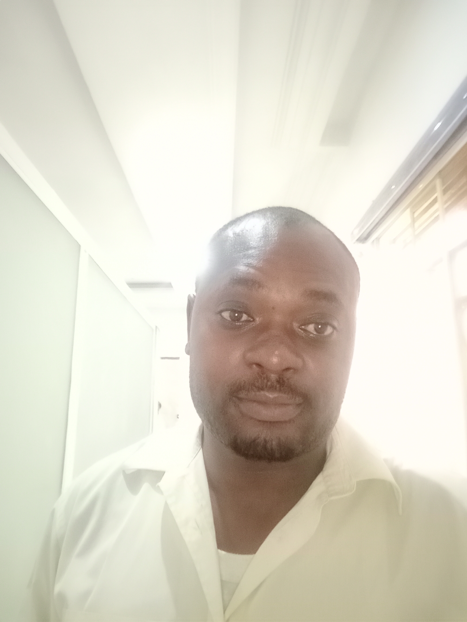 The image shows a head-and-shoulders self-portrait of a man with dark skin, a shaved head, and a short beard. He is wearing a light-colored collared shirt, possibly white. The background appears to be an indoor space, possibly a hallway or office, with light-colored walls and a white ceiling with recessed lighting. The location is described as Gwarinpa, Nigeria, but there are no specific details in the image to visually confirm the city.