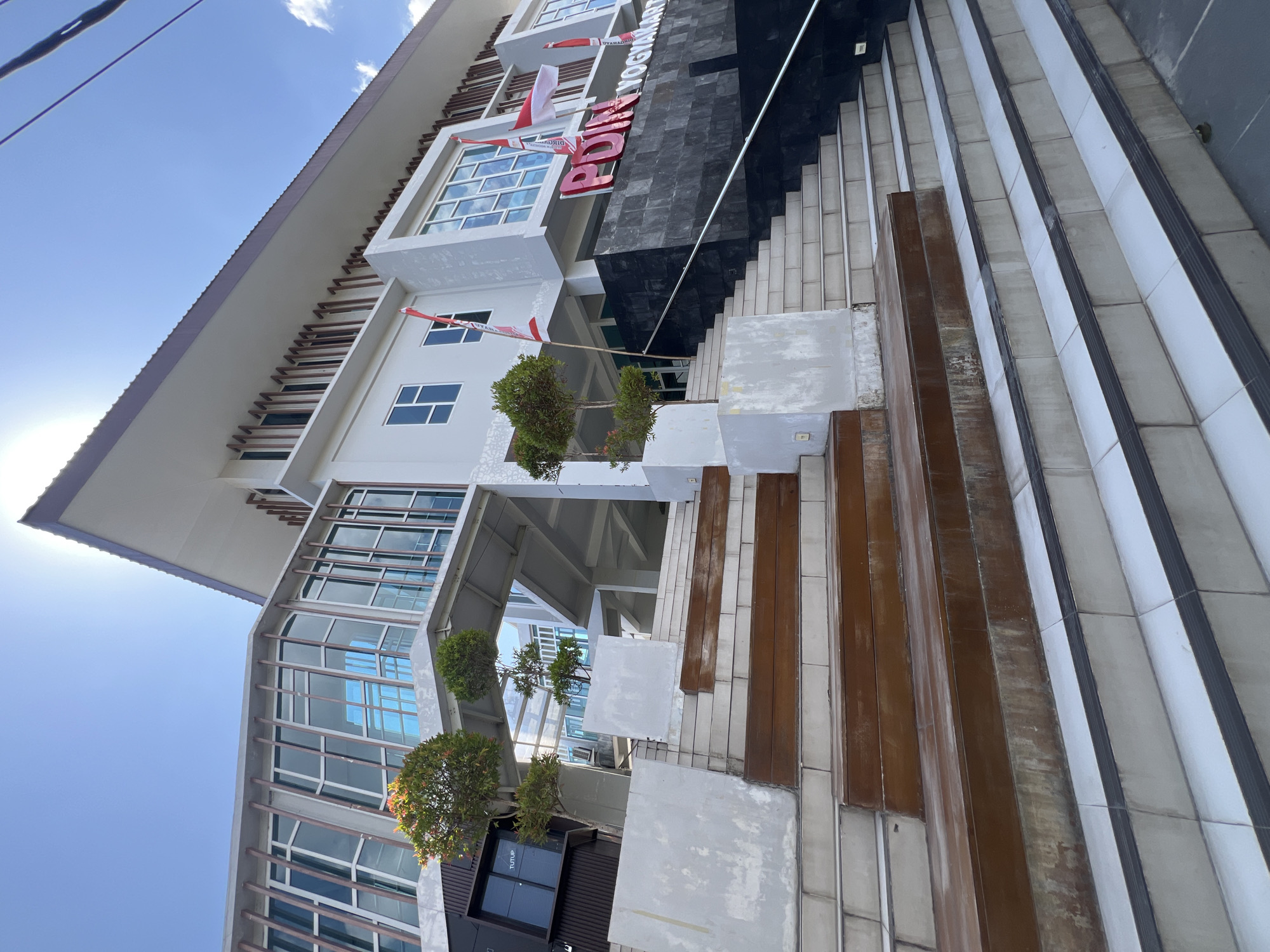 The image shows the exterior of a modern building in Yogyakarta, Indonesia, with a wide set of stairs leading up to the entrance. The building features a contemporary design with large windows, a light-colored facade, and dark stone accents. Several Indonesian flags are visible on the building, indicating a national or ceremonial context. The stairs are a prominent feature, constructed with both concrete and wooden elements, some of which appear to be weathered. Trees are planted on landings along the stairs, adding a touch of greenery to the scene. The sky is bright blue with some clouds, suggesting a sunny day. The building appears to be an institutional or public building, possibly a government office or educational facility, given the prominent signage that reads "PDIN YOGYAKARTA".