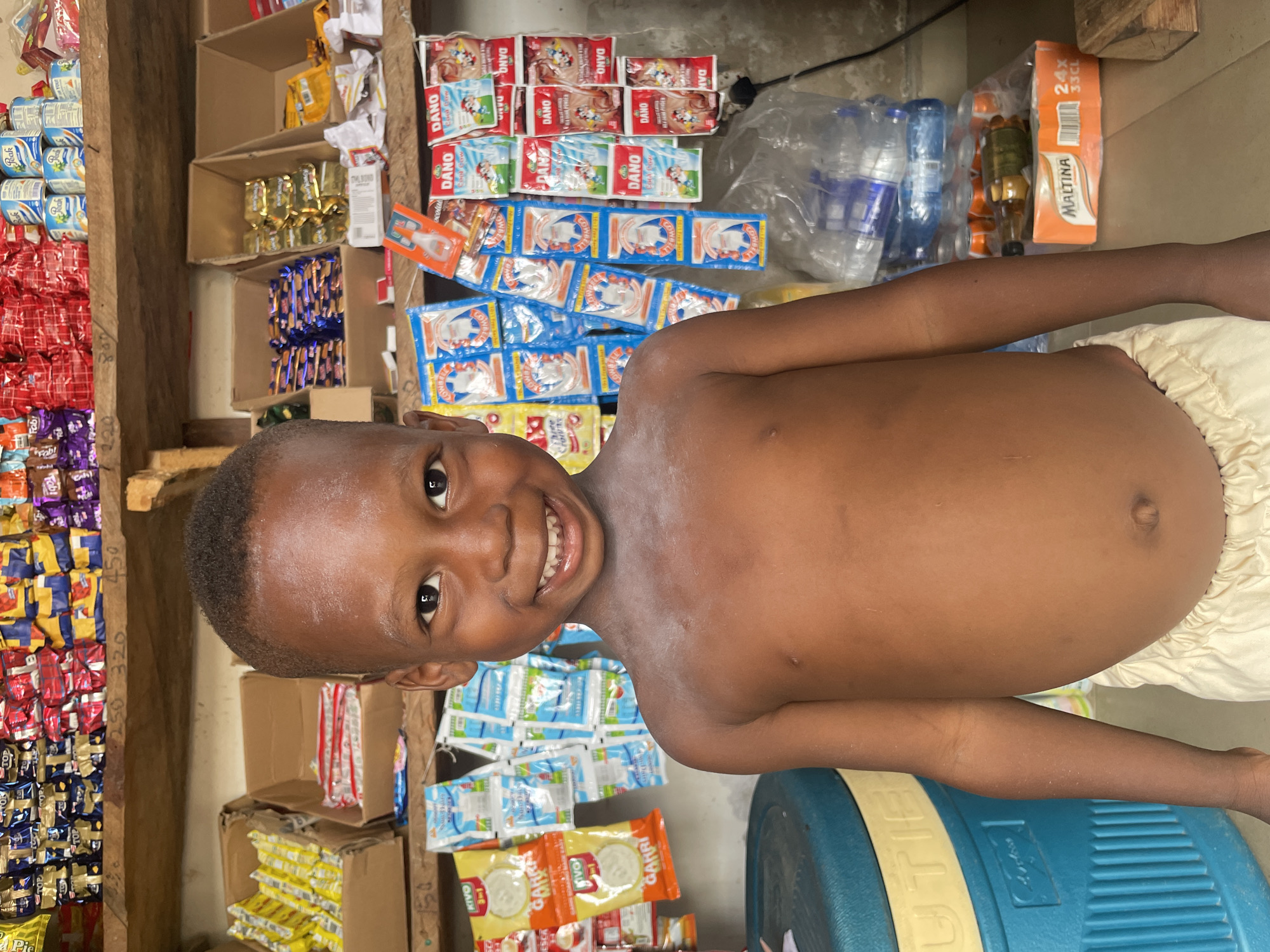 A cheerful young child, appearing to be a boy, is the central subject of this portrait taken indoors. He is shirtless and wears light-colored shorts or a diaper, smiling broadly and looking directly at the camera, conveying a sense of happiness.
The background reveals a bustling small convenience store or market stall, typical of a local shop in Lokoja, Nigeria. Products are densely packed on simple wooden and cardboard box shelves. Visible items include numerous sachets of "DANO" milk powder (in both red and blue packaging) and "Peak" milk cans. Various packaged snacks or candies in red, purple, blue, and yellow wrappers, some labeled "TOP," are stacked. Also visible are "KIVO" and "GANRI" branded items in yellow and orange sachets.
Bottles of water and a carton of "MALTINA" (marked "24X 33CL") are visible towards the upper right. Small plastic-wrapped items, including what appears to be a syringe or similar tool in orange packaging, are also on display. A blue and yellow cooler or container is partially seen in the lower right foreground. Price markings like "320" and "450" are faintly visible on some shelves. The setting suggests a modest, well-stocked local establishment, with the child playfully interrupting the scene. The lighting is consistent with an indoor setting, likely during the daytime.