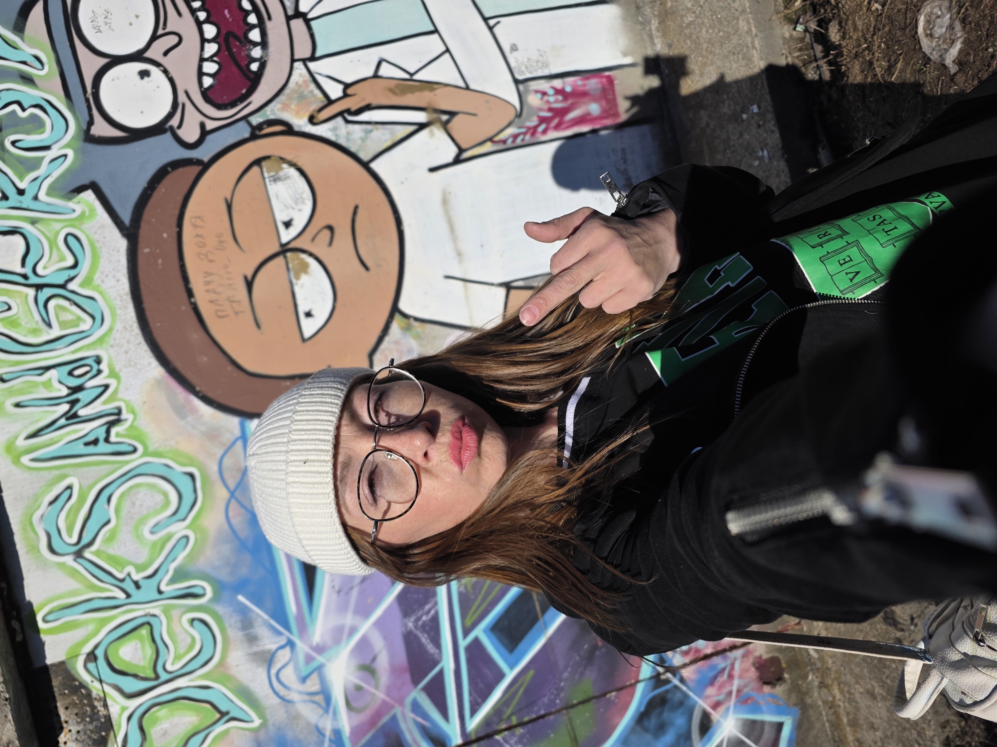 A woman wearing a white beanie and round glasses is taking a selfie outdoors in front of graffiti art featuring characters from the Rick and Morty cartoon. The woman points at the graffiti with her left hand. She is wearing a black jacket with a green Harvard logo visible. Part of a white sneaker is visible in the lower right corner. The setting is in Chaikovsky, Russia.