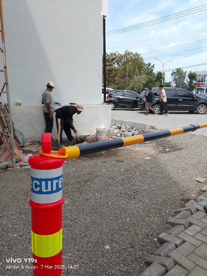 The image shows a scene in Indramayu, Indonesia, at the coordinates -6.343 latitude and 108.335 longitude. Three men are working on the construction of a new gate. The men are wearing casual clothing, including one man wearing a hat. One man is using a shovel to move dirt and gravel, while the others are standing by. The gate is a black and yellow painted barrier that is partially constructed and is laying on the ground in front of a white wall. In the background, there is a black car parked on a paved road. The pavement around the barrier is made of small gravel and loose stones. There is a large, red and yellow, traffic cone marked "secure" standing in the foreground of the image.