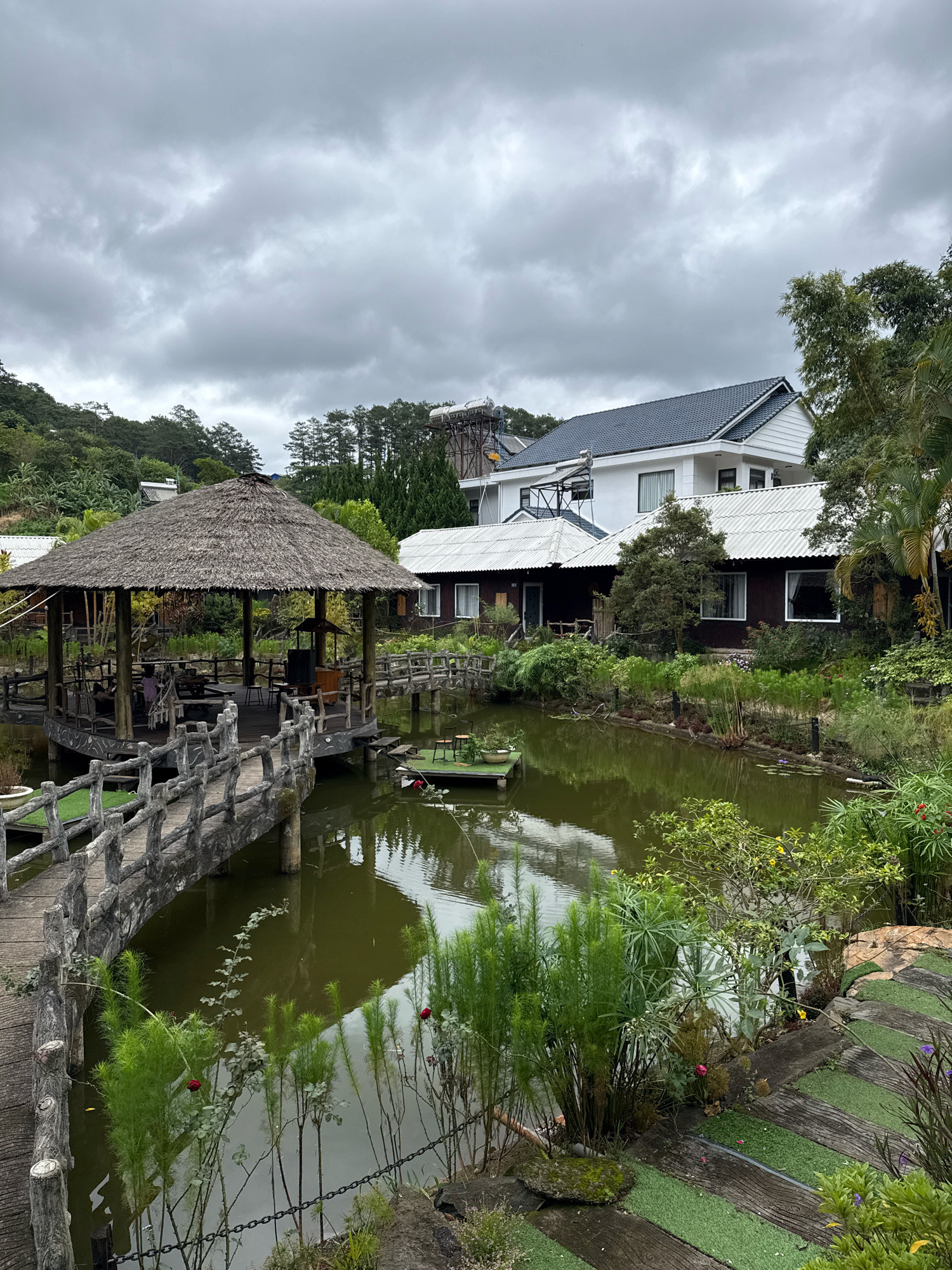 The image captures a serene scene at a resort or guesthouse in Hiệp Thạnh, Vietnam. The focal point is a thatched-roof pavilion situated on a wooden platform over a calm, greenish pond. A wooden bridge with a rustic, concrete railing leads to the pavilion. The pavilion appears to be an outdoor seating or dining area, with tables and chairs visible. Surrounding the pond is lush greenery, including trees, bushes, and ornamental plants. In the background, a modern white building with a dark tiled roof stands out, contrasting with the natural elements. The sky is overcast with gray clouds. There are no people clearly visible engaging in any actions. The overall impression is one of tranquility and a harmonious blend of natural and built environments, typical of a tourist destination.