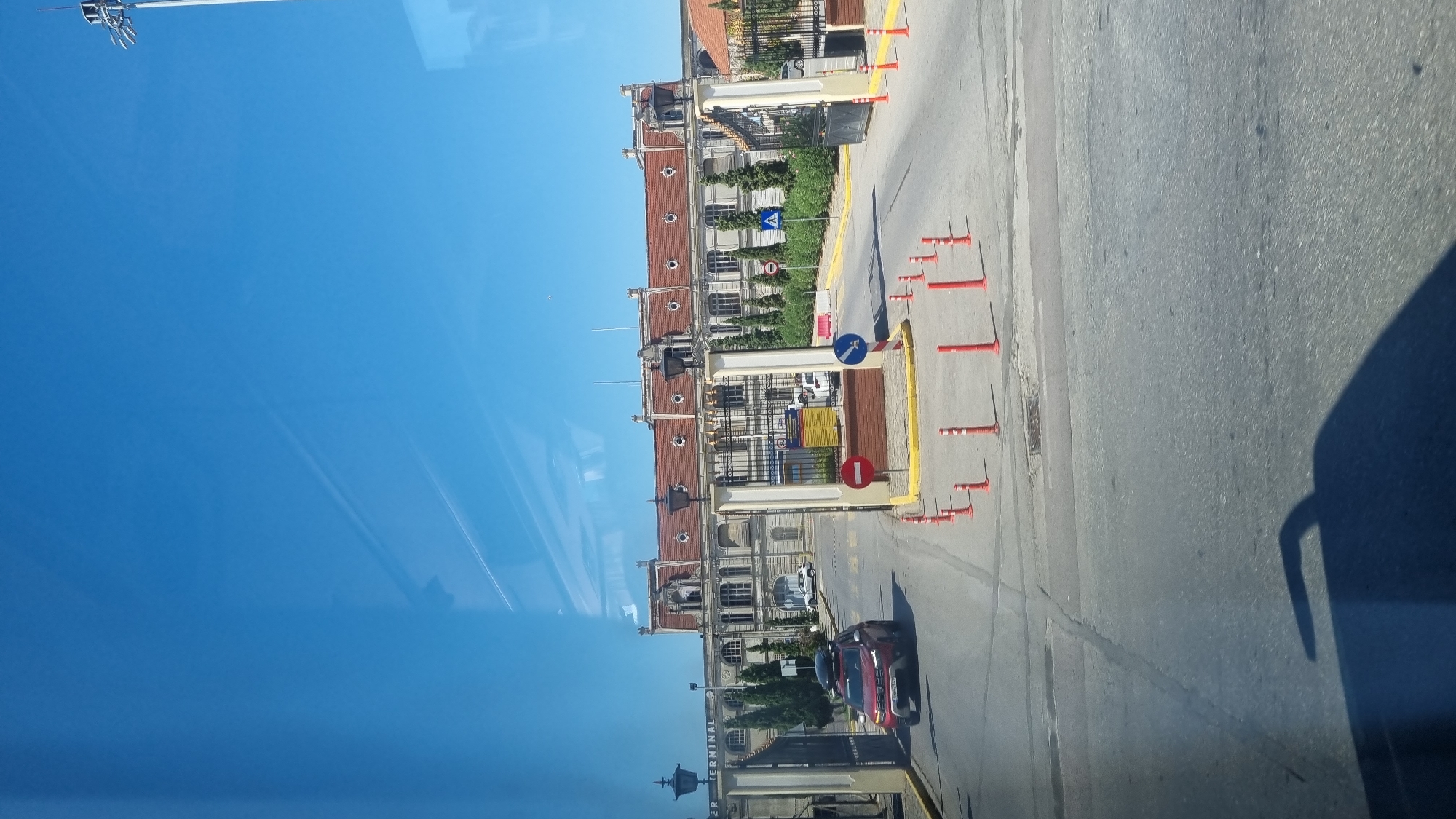 The image shows a red SUV entering a gateway. The gateway leads to a large, classical-style building with a red tiled roof. The building appears to be a terminal of some kind, with the word "TERMINAL" visible on the left side of the gateway. The scene is set on a sunny day, with a clear blue sky. The area in front of the building is paved, with several orange traffic cones and bollards lining the road. The overall impression is that of a transport hub or a public institution.