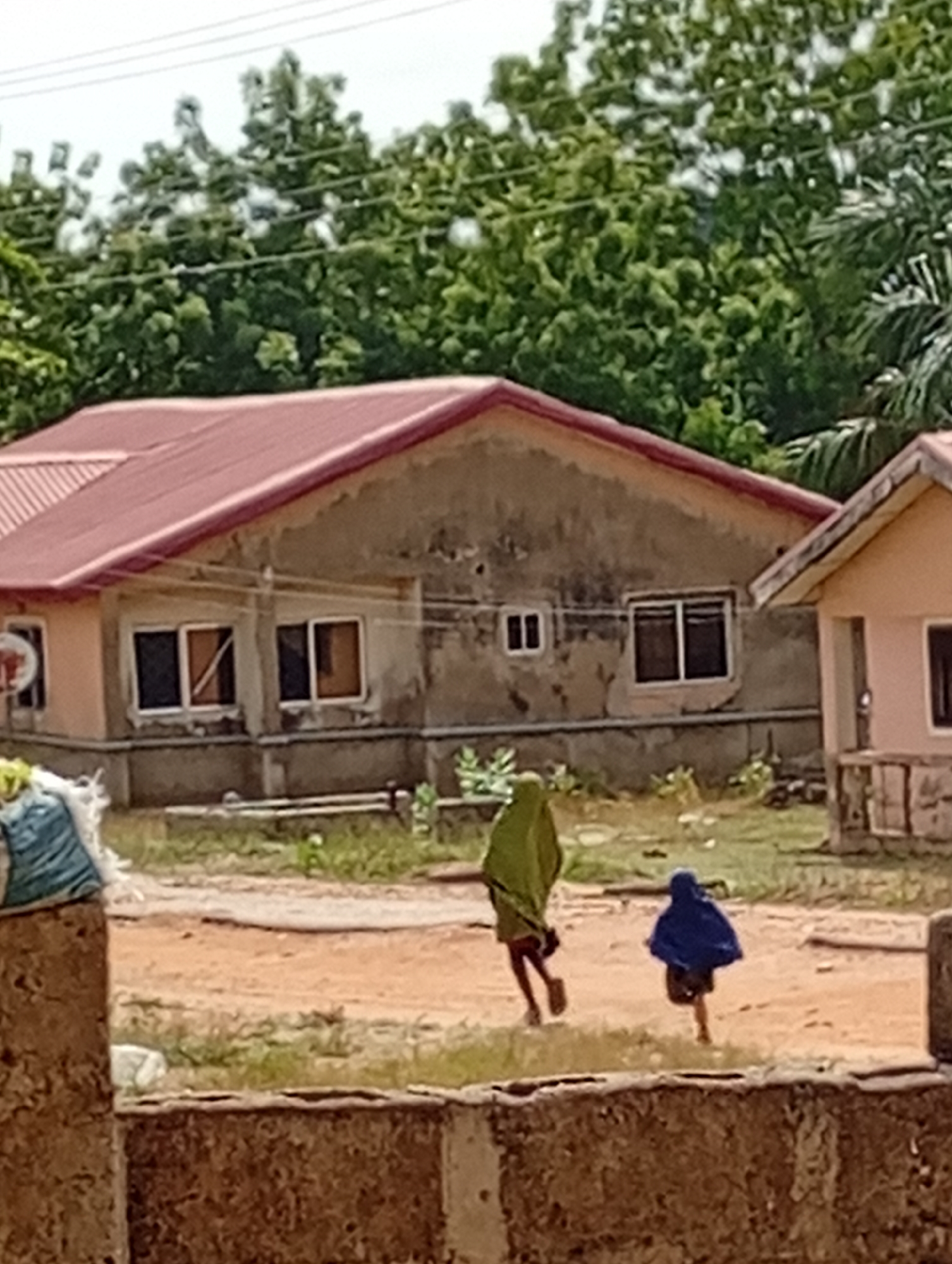 Two people are running on a dirt field in front of a building. The person on the left is wearing a green robe and the person on the right is wearing a blue robe. The building is a single-story structure with a red roof. There are trees in the background. The location is Unknown, Unknown.