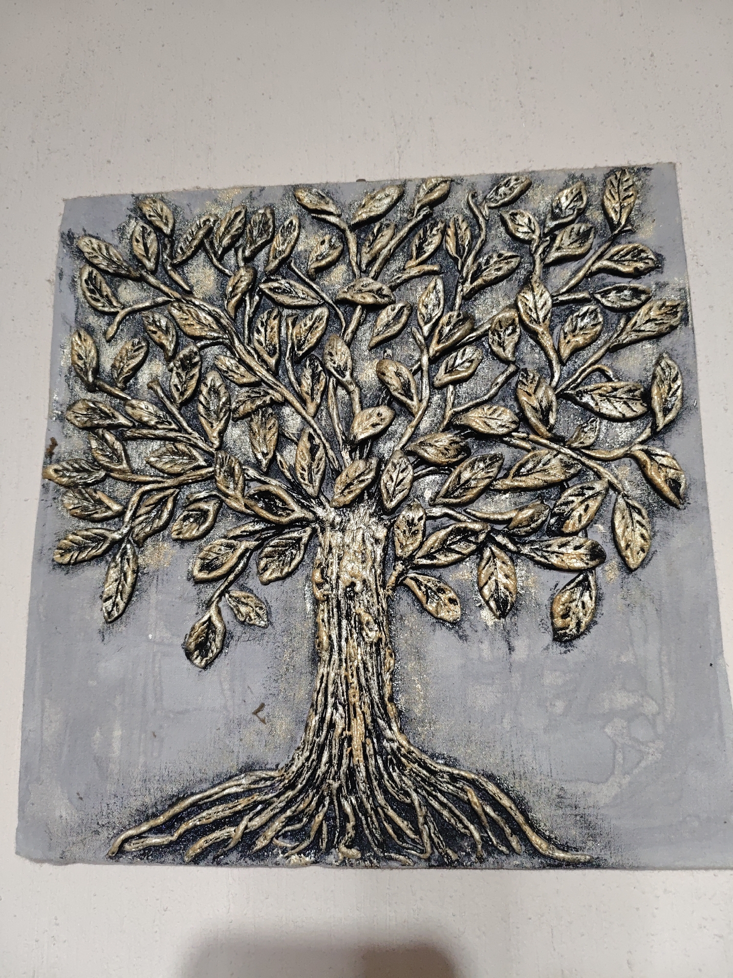 The image depicts a square, raised-relief wall art piece featuring a stylized tree with roots, trunk, branches, and leaves. The tree is primarily black with gold highlighting. The background of the artwork is grey with similar gold accents. The piece is mounted on a white wall.