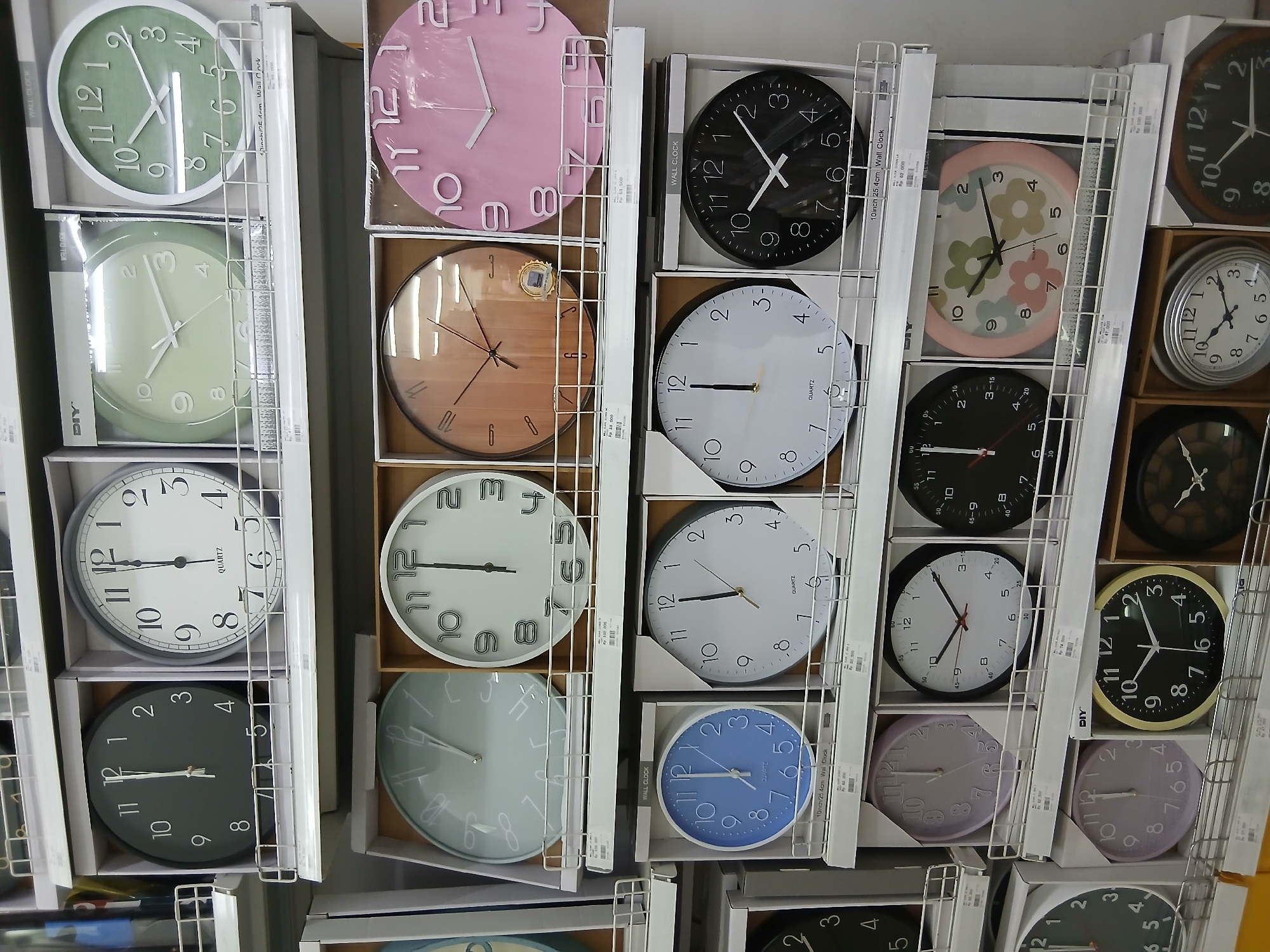This image displays a retail shelf filled with various wall clocks. The clocks are presented in their original packaging and are arranged in rows on white shelving units. The clocks vary in color, style, and size, with some featuring Roman numerals and others Arabic numerals. The scene appears to be an indoor retail environment, likely a home goods or department store. The presence of price tags suggests the items are for sale. This is a close-up shot, focusing on the merchandise. The environment is well-lit, with fluorescent lighting typical of a commercial space. No people are visible in the frame. There is no specific indication of location within South Jakarta, Indonesia, as the image focuses solely on the product display.