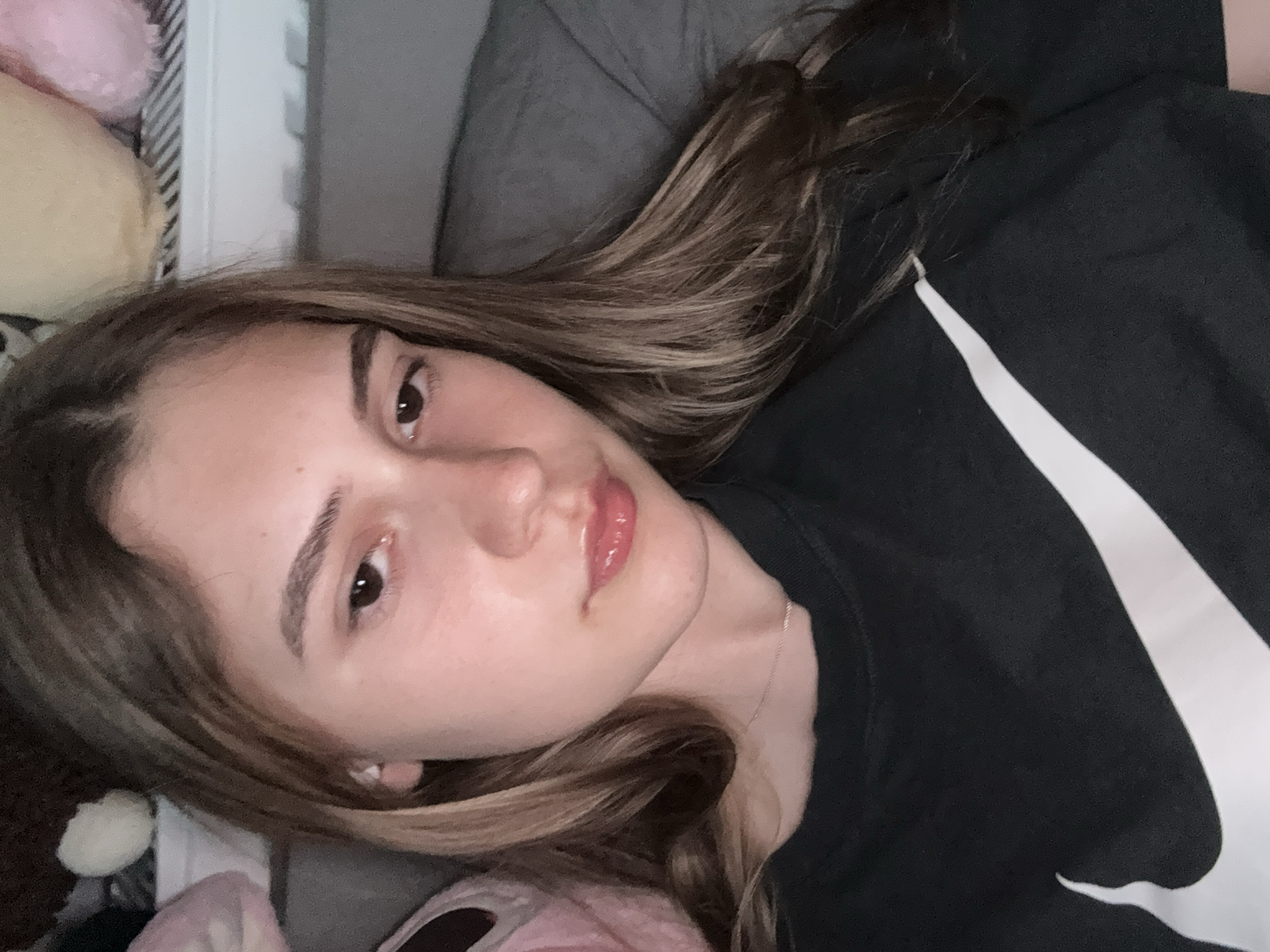 A young woman with light brown hair and brown eyes is lying down, looking directly at the camera. She is wearing a black Nike shirt with a white swoosh logo. Her face is clear, with a slight sheen on her lips. Behind her are soft, pastel-colored objects, potentially plush toys, and a gray surface, likely a pillow or bedding. The scene is indoors. There is no indication of the city of Panagyurishte, Bulgaria in the provided image.