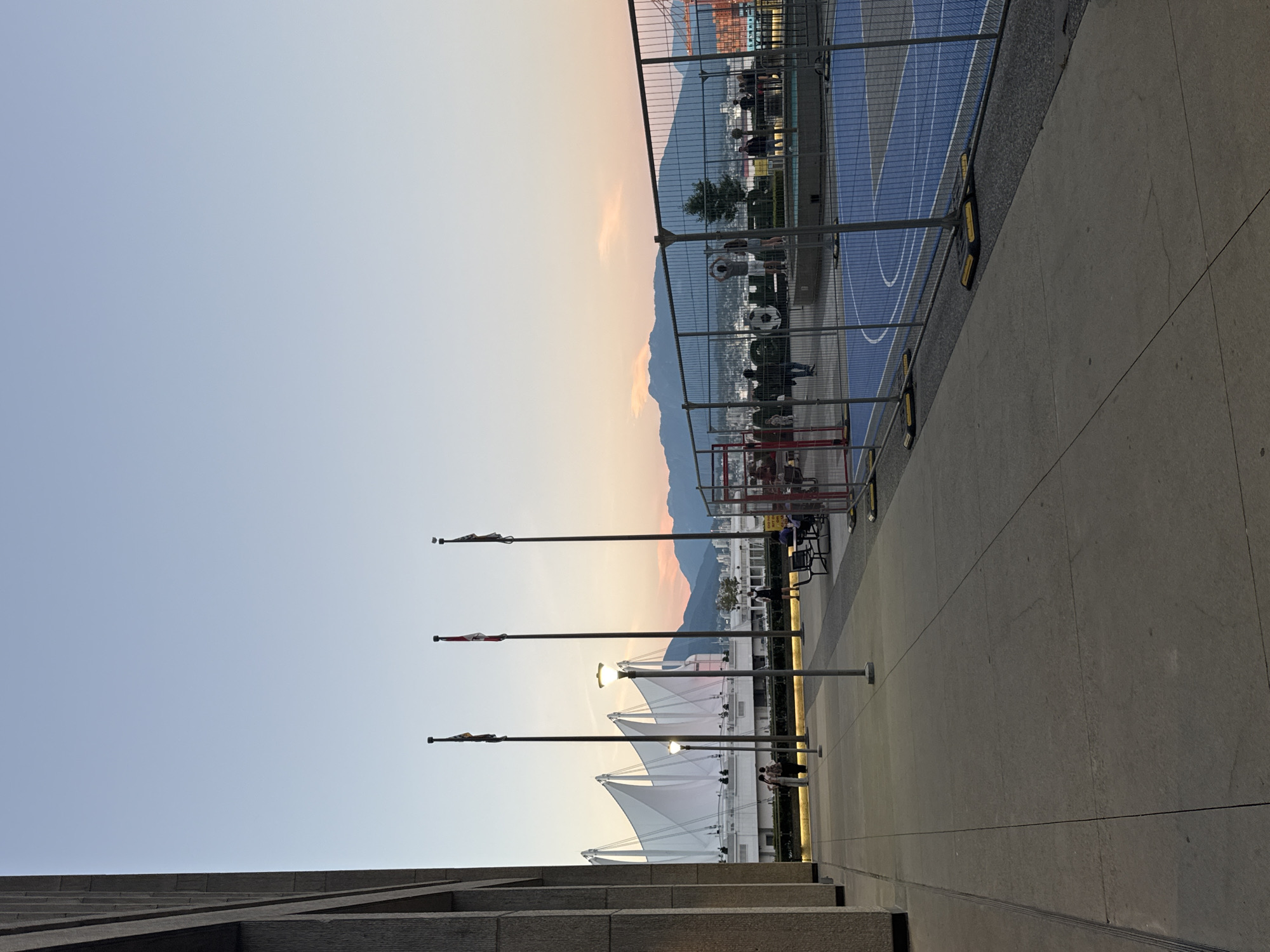 An outdoor shot captures a sunset scene in Vancouver, Canada, with a view of the mountains in the background. In the foreground, a concrete sidewalk runs parallel to a building's stone facade on the left. To the right, a fenced-off blue court, possibly for basketball, is visible. Several people are scattered across the scene: two individuals are conversing near a bench in the mid-ground, while a few others are seen near the court and in the distance. Three flagpoles with flags stand tall, and illuminated streetlights dot the area. The iconic white sails of Canada Place are a prominent landmark in the background, adding to the city's distinctive skyline. The sky displays a gradient of light blue to soft orange hues, indicative of twilight.
