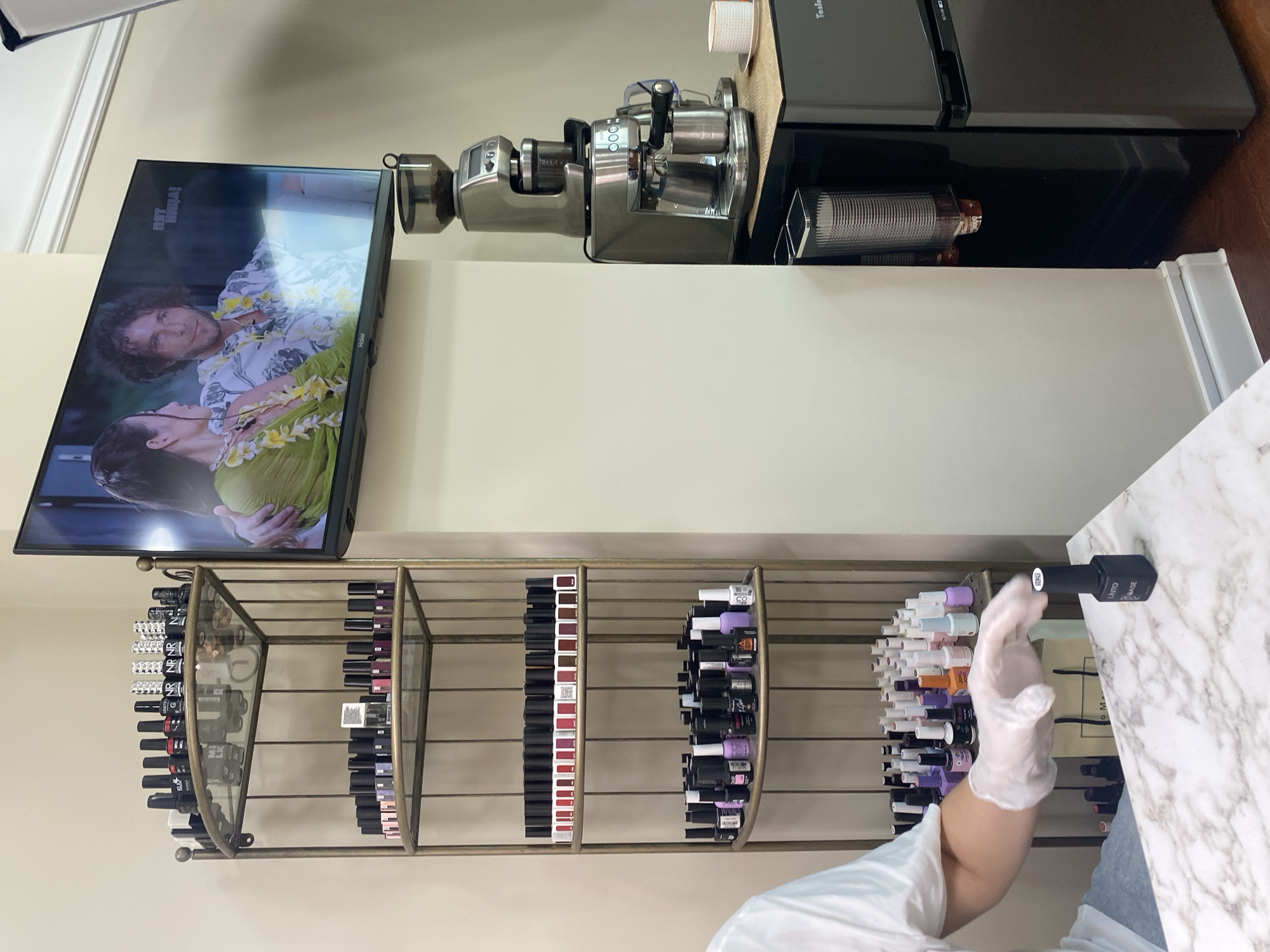 The image shows an interior scene with a television mounted on a light-colored wall, displaying an image of a couple. Below the television, there's a metallic shelf unit filled with nail polish bottles of various colors. To the right of the television, there's a metallic appliance, possibly a coffee maker, sitting atop a black mini-refrigerator. A person wearing white gloves and a white shirt is holding a nail polish bottle near a white marble-patterned countertop. The environment appears to be a beauty salon or a similar workspace. There's no direct indication of the city of Sochi or any other location details.