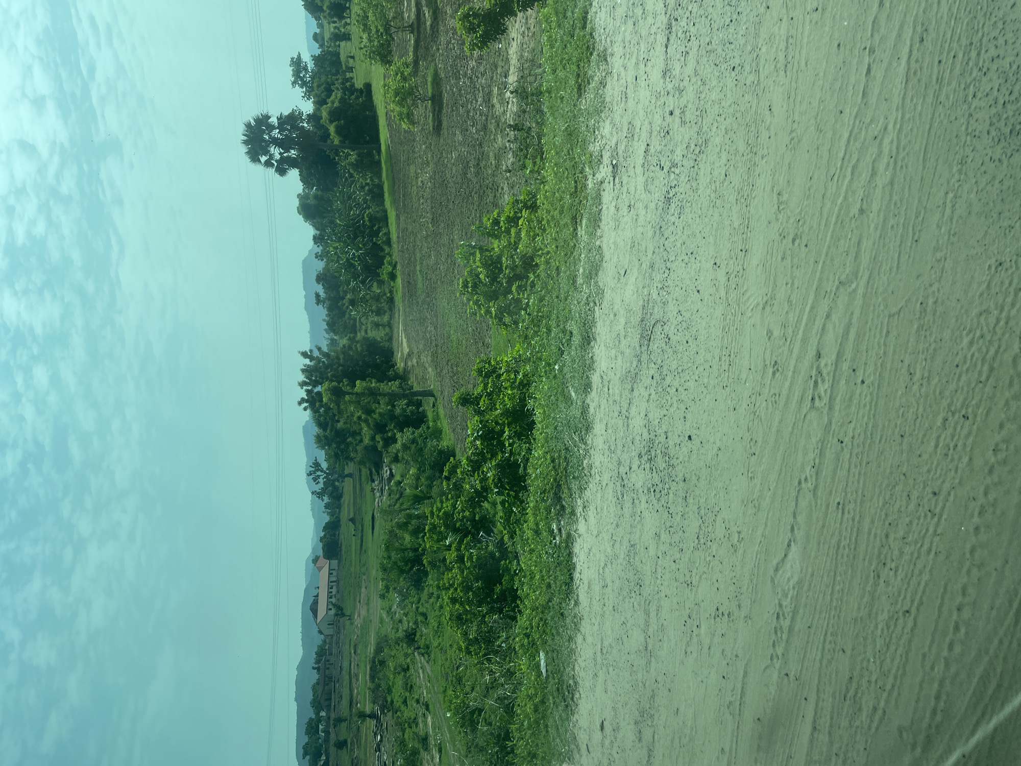 The image depicts a rural landscape in Chum, Nigeria. In the foreground, a sandy path with tire tracks is visible. Beyond the path, lush green vegetation, including trees and bushes, dominates the scene. In the mid-ground, a cultivated field with dark soil contrasts with the surrounding greenery. A few scattered trees are also present in this area. Further in the background, several buildings are visible, including what appears to be a church or school with a red roof. Rolling hills and mountains form the backdrop, with a hazy sky overhead. The overall impression is one of a tranquil, rural setting in the Nigerian countryside.