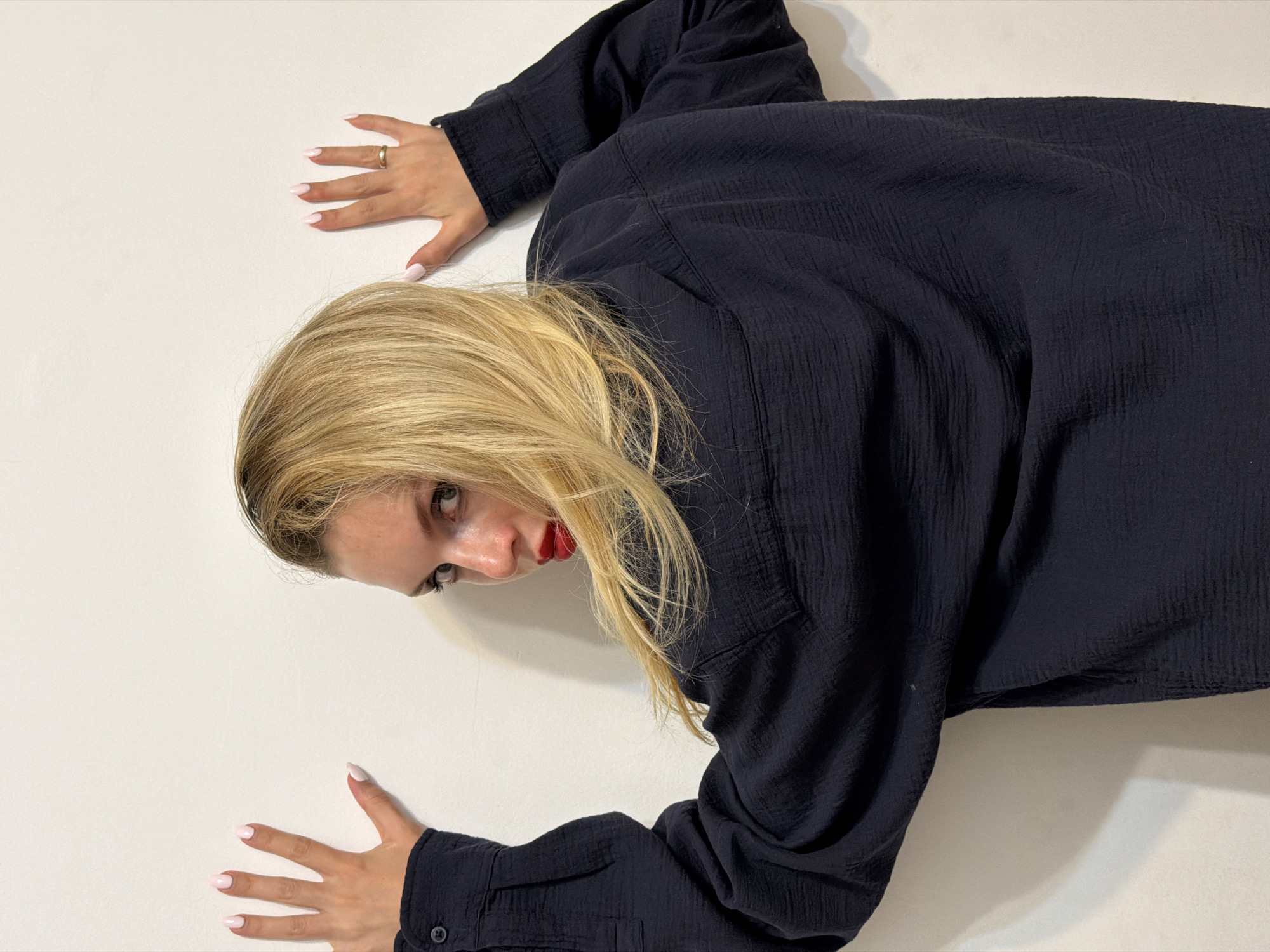 A woman is lying on a white surface. She has long, blonde hair and is wearing a dark, long-sleeved shirt. Her hands are positioned as though she is holding onto the surface, with pale pink nail polish visible. The surface is likely a wall. The scene's location is not discernable from the image.