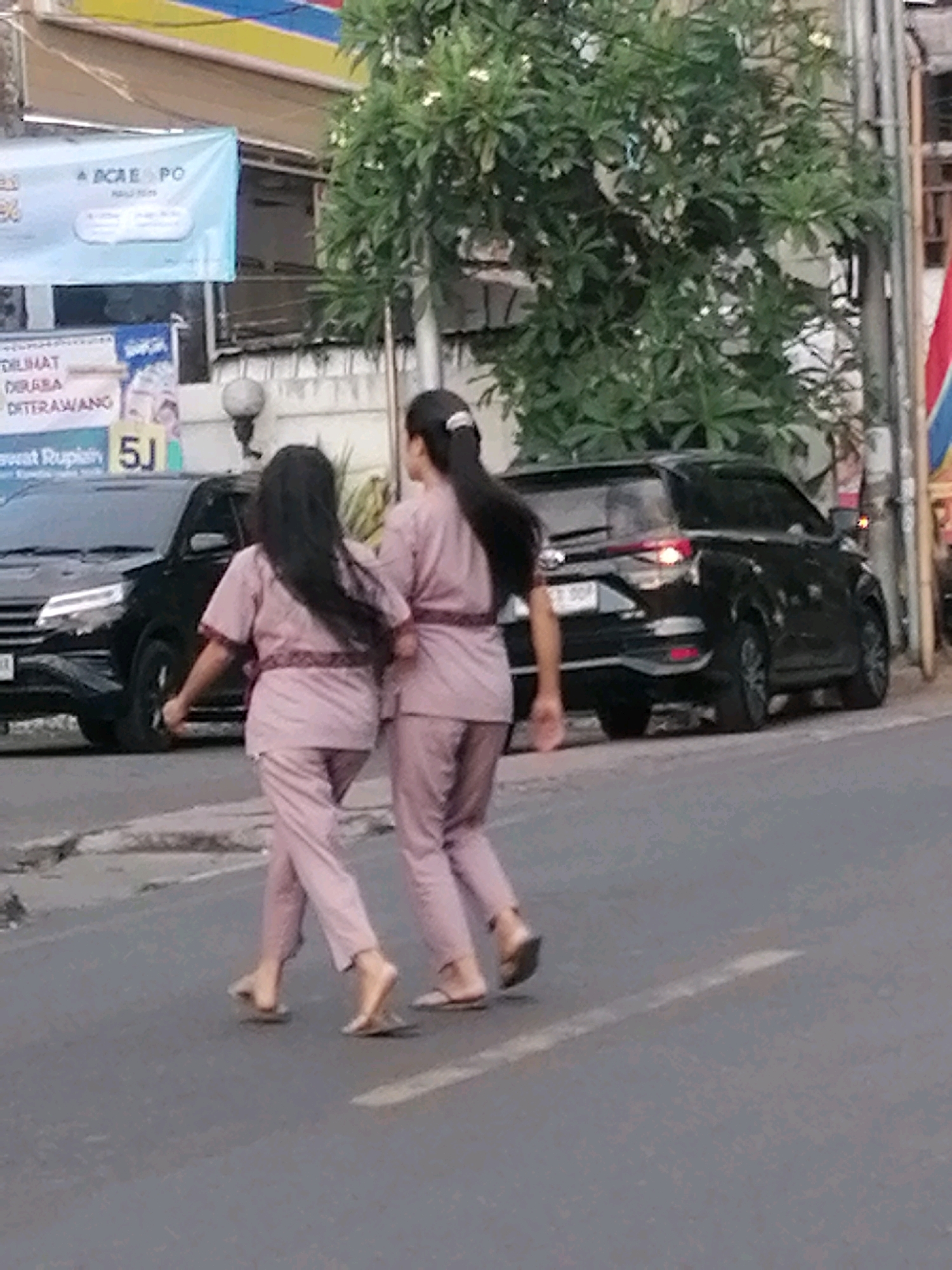 Two young women wearing matching pink uniforms with dark trim are walking across a street in a city. The woman on the left has long black hair and is walking barefoot. The woman on the right has her hair in a ponytail and is wearing sandals. A black SUV is parked on the side of the road behind them. In the background, there is a building with banners and trees. The scene appears to be in Ubud District, Indonesia, given the presence of local signage and vegetation. The lighting suggests it is daytime.
