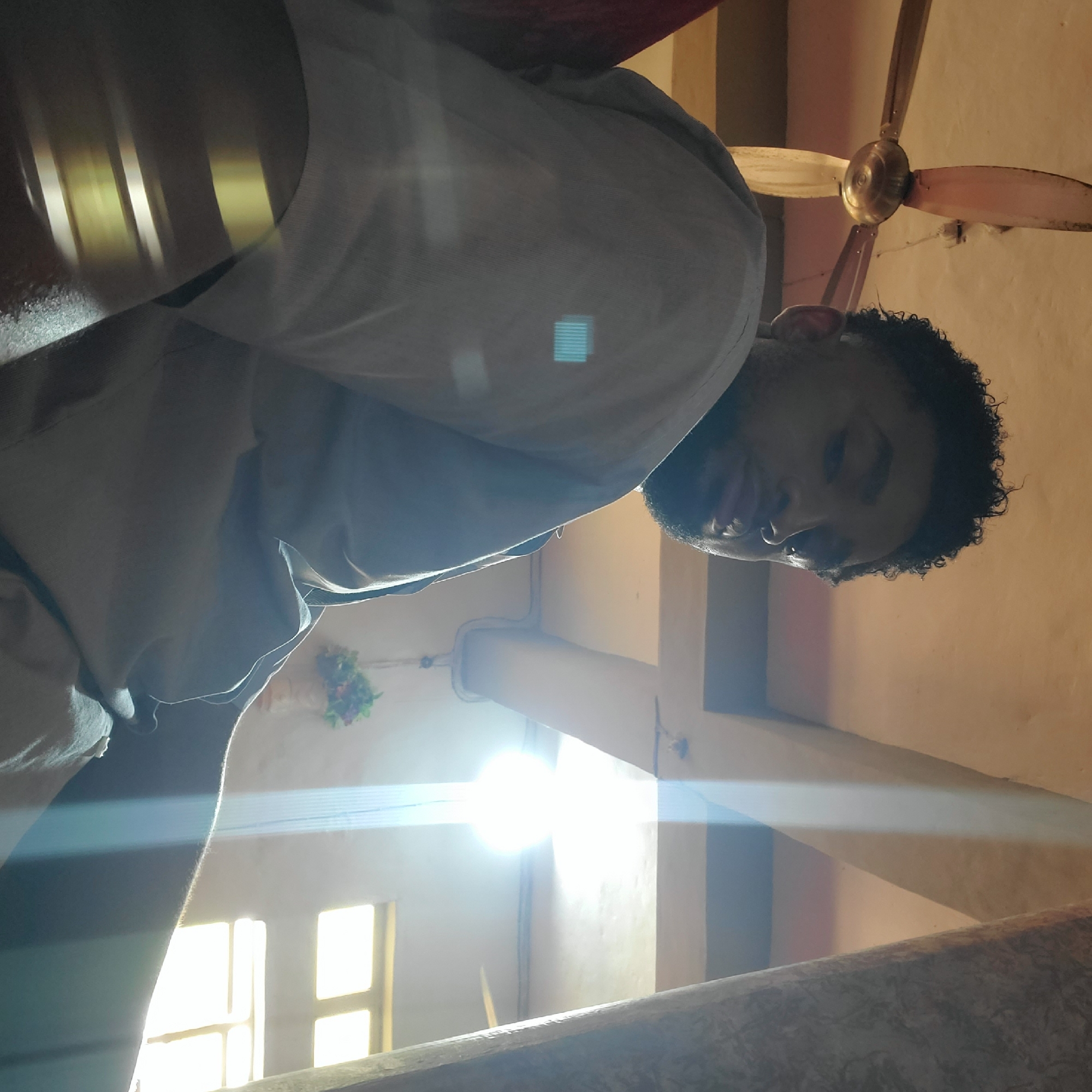 A man with curly dark hair and a nose ring looks downward towards the camera. He wears a light-colored shirt. He appears to be in a building with tan walls, a white ceiling fan, and a window with sunlight streaming in. There is a floral arrangement attached to a wall. The location appears to be Onitsha, Nigeria.
