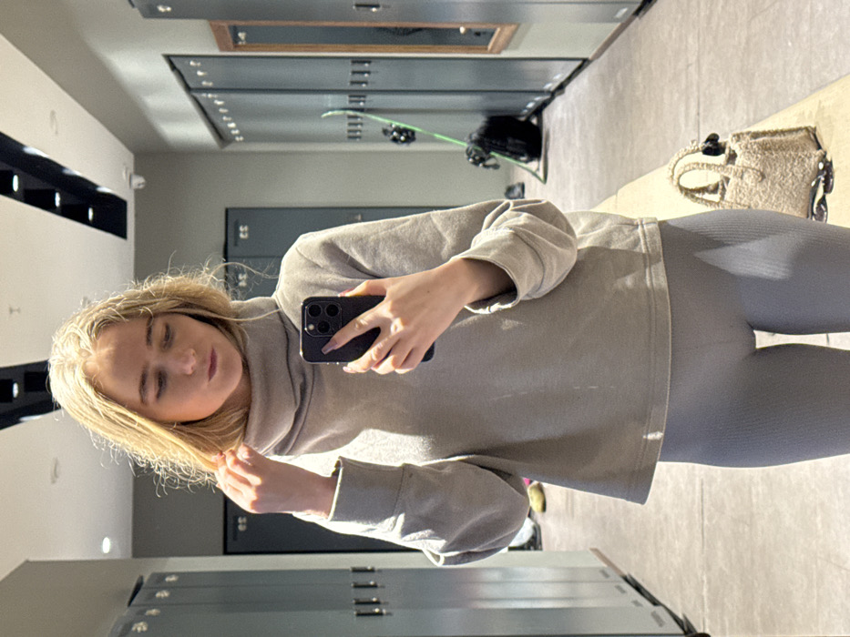 A blonde woman in grey leggings and a matching sweatshirt takes a selfie in a locker room. Lockers line the walls. Ski equipment is on top of the lockers across from her. A light-colored bag sits on the floor. She is holding a dark-colored smartphone with three prominent lenses.