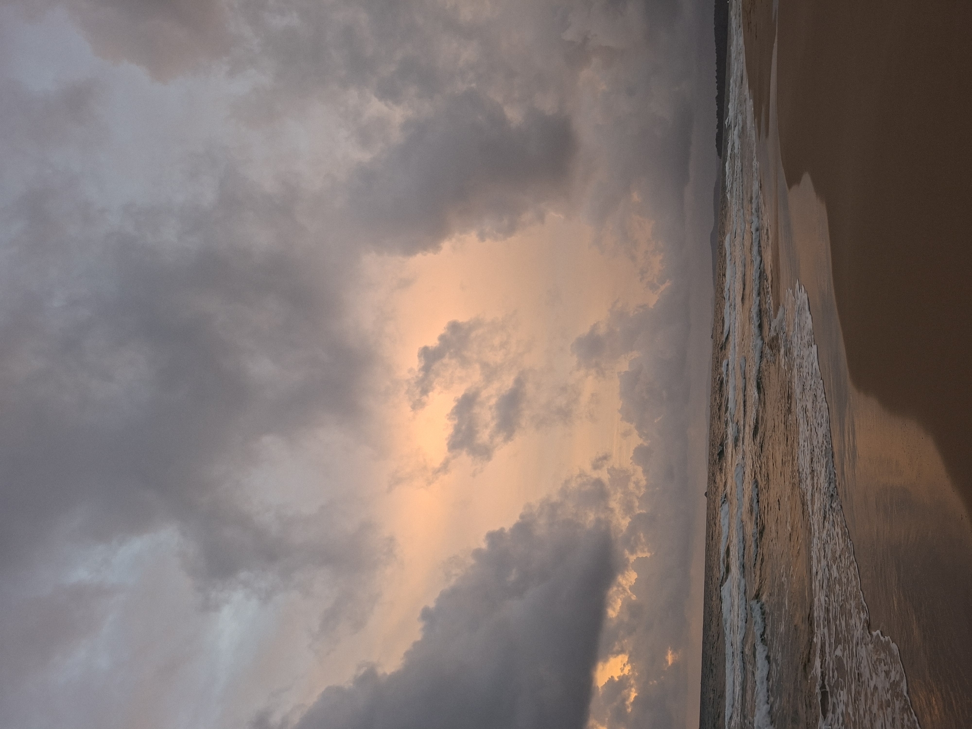 A serene beach scene unfolds under a dramatic sky, likely captured at sunset in Phú Quốc, Vietnam. The sky is a canvas of swirling, heavy clouds, tinged with a soft peachy glow peeking through the center, suggesting the fading sun. Darker, voluminous clouds dominate the upper and side portions of the frame. Below, the ocean meets a sandy shore with gentle waves lapping at the wet sand. The water is a muted, reflective hue, mirroring the sky's colors. The beach itself is a smooth expanse of sand, with the shadow of an unseen object stretching across it from the right. In the distance, a faint outline of landmass and trees suggests the coastline of Phú Quốc. No people or discernible objects are present on the beach or in the water, emphasizing the natural and tranquil atmosphere of the location.