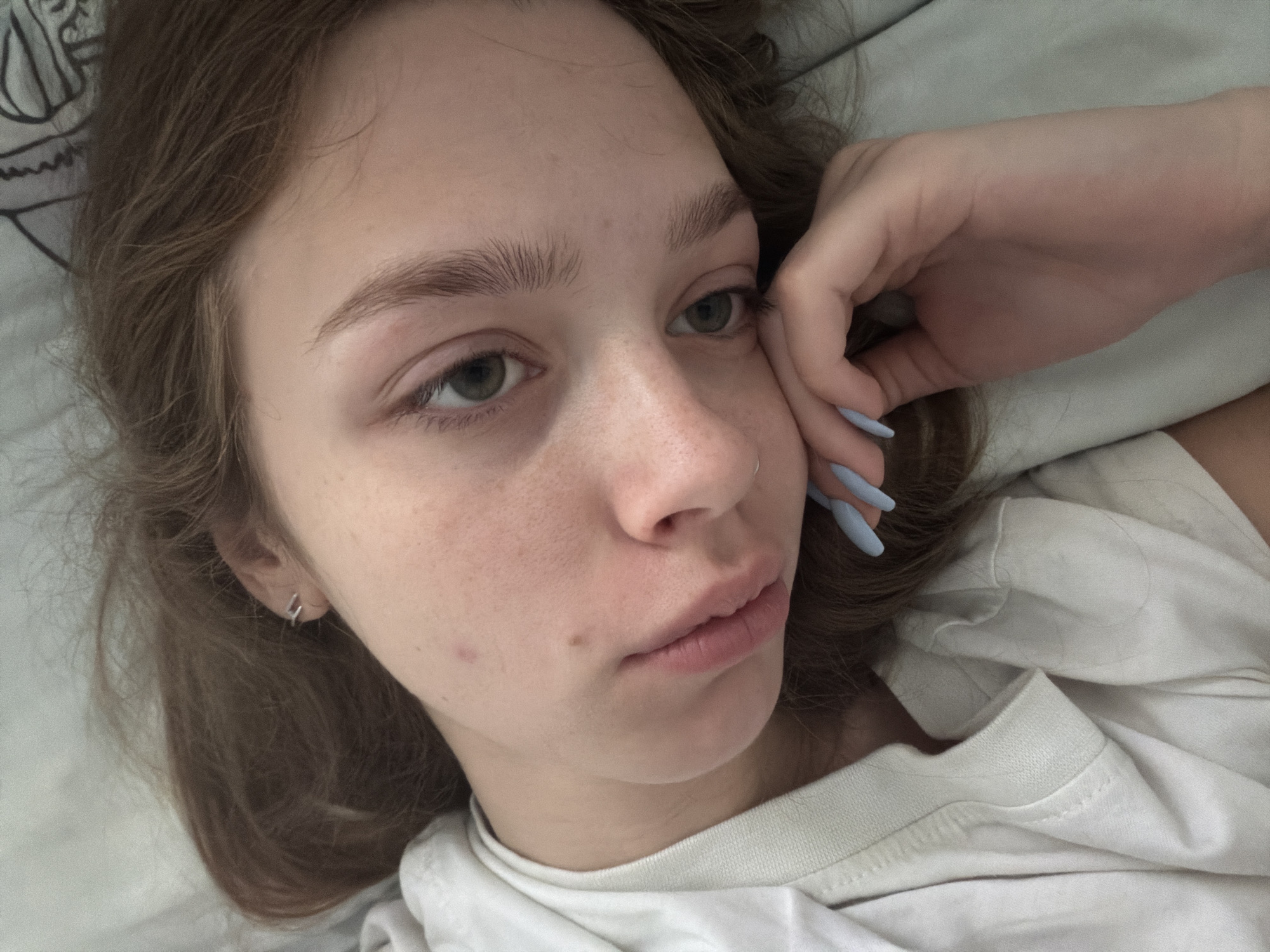 A young woman with light brown hair lies on her side on a bed. She is wearing a white t-shirt. Her right hand is resting on her face. Her fingernails are painted a light blue color. The background is a light blue comforter. The scene appears to be indoors, likely in a bedroom, in an unknown location.