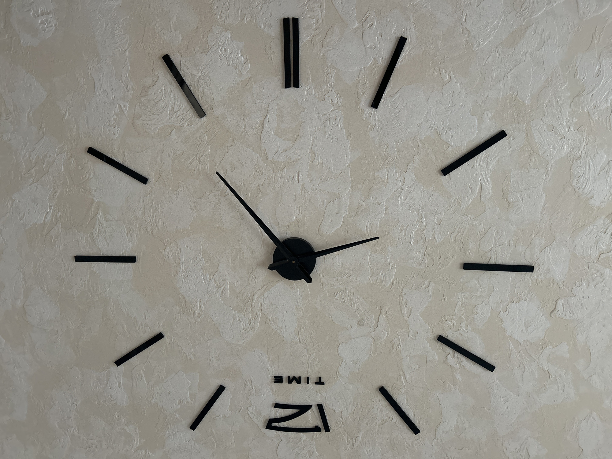The image shows a modern, minimalist wall clock mounted on a textured light-colored wall. The clock has no numerals, only black hour markers in the form of short, thick lines. The word "TIME" is printed in black capital letters below the "12" marker. The clock hands are also black and appear to be made of metal. The hour hand is pointing slightly past the "10", and the minute hand is pointing directly at the "4". The image is a close-up of the clock and does not provide any environmental context beyond the wall it is mounted on. There are no people or other objects visible in the image. The location in the city of Uvel'skiy, Russia is not determinable from the image itself.