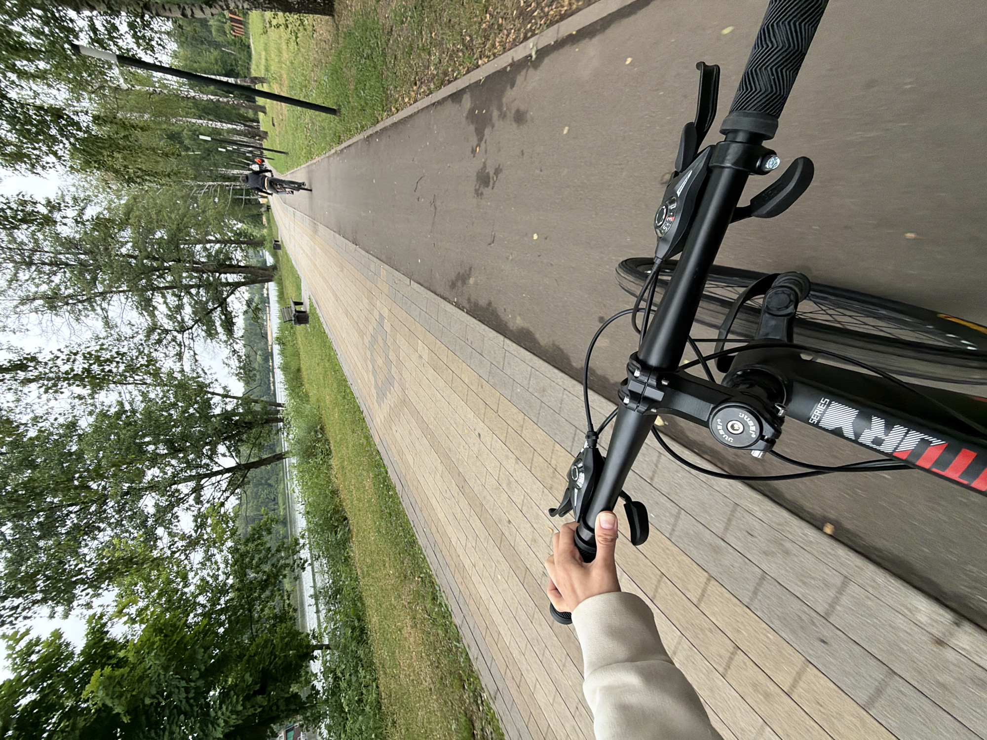 A first-person perspective shot shows a person riding a bicycle on a paved path in Malakhovka, Russia. The path runs alongside a body of water with trees and greenery on the opposite bank. The foreground is dominated by the bicycle's handlebars and the rider's hand. The path itself is a mix of grey asphalt and lighter-colored paving stones. In the distance, another cyclist is visible on the same path. The sky is overcast.