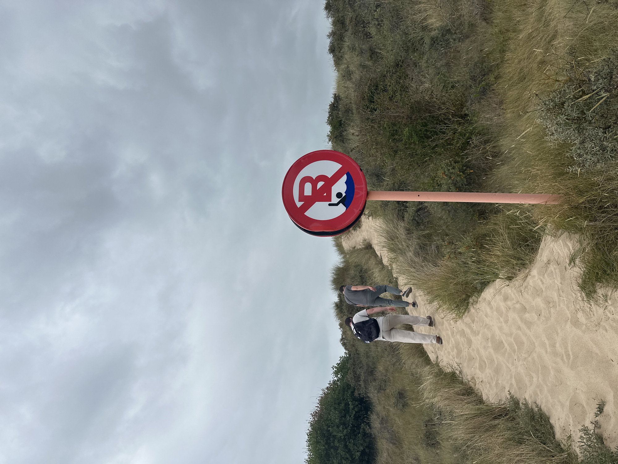 A sign depicting a person in water with a red circle and a diagonal line through it is displayed in the foreground of the image. The sign is mounted on a thin orange pole. Behind the sign, two individuals are walking up a sandy path that winds through a dune landscape. The path is bordered by tall grasses and sparse vegetation. The sky above is overcast with a gray, cloudy appearance. The scene is set in the natural dune environment of Knokke-Heist, Belgium, likely along the coastline.