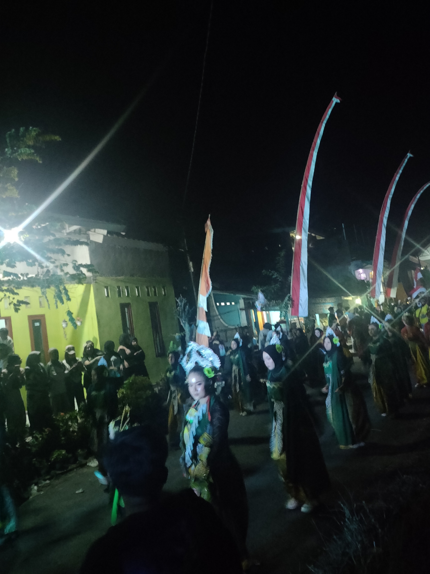 In Tongaswetan, Indonesia, at night, a parade is underway. The participants, adorned in traditional attire and elaborate headdresses, move along a street lined with buildings. Several individuals, possibly performers, are dressed in vibrant green and yellow costumes. Tall, red and white striped flags, likely representing national or local symbols, are erected along the procession route. The scene is illuminated by artificial lights, casting shadows and highlighting the festive atmosphere. A crowd of onlookers, some in darker clothing, observes the parade from the sides of the street. The architecture of the buildings appears to be modest, with some featuring windows and doorways. The ground is paved, indicating an urban or semi-urban setting.