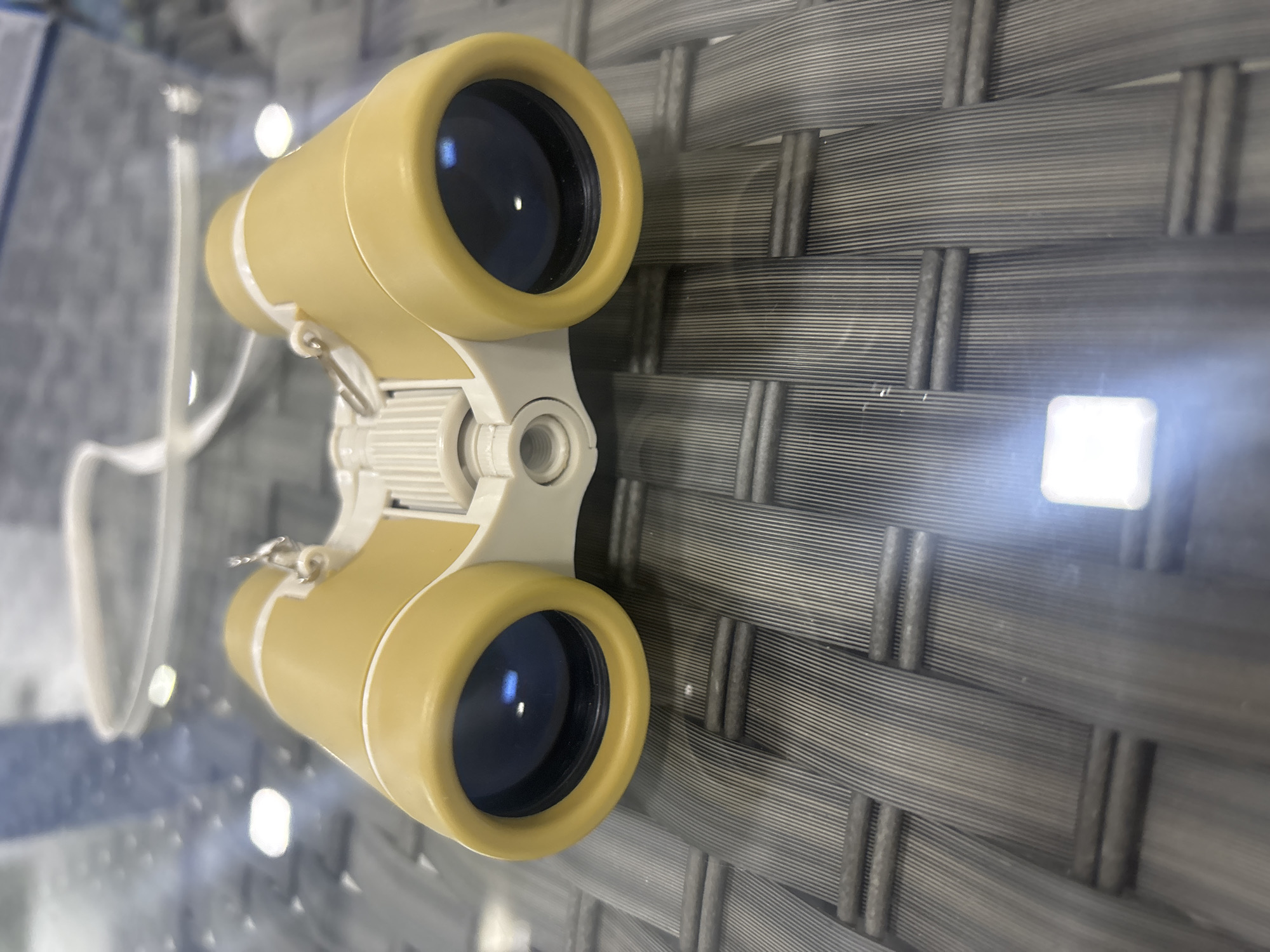 A pair of yellow and white binoculars lies on a dark gray woven surface, possibly a table. The binoculars are positioned front-facing, with the eyepieces visible. A white strap is attached to the binoculars and arcs upwards and out of frame. The surface beneath the binoculars shows a distinct woven pattern with alternating dark and lighter gray strands. There is a bright, rectangular reflection on the surface towards the bottom center of the image. The background is blurred and appears to be a combination of glass surfaces and possibly vertical structures. No people are present in the image. The scene is indoors. The location is not specified.