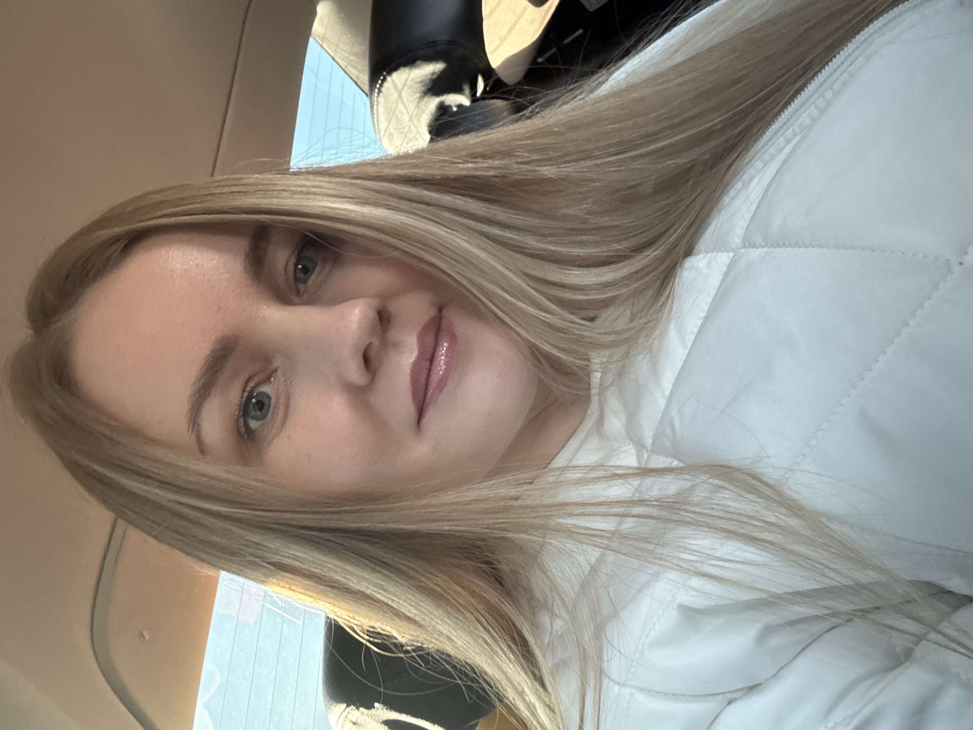 A fair-skinned woman with long blonde hair is sitting in what appears to be a car. She is wearing a white quilted jacket. The background shows a window and a glimpse of the car's interior. The location within Chaikovsky, Russia, cannot be determined from the image.