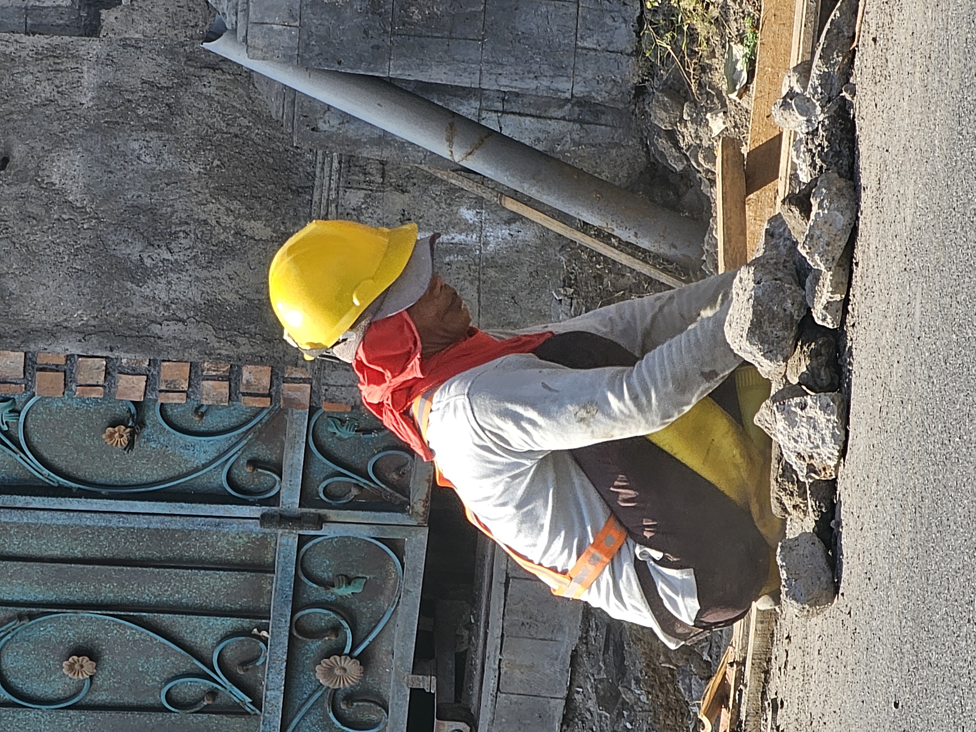 A person wearing a yellow hard hat, a red cloth around their neck, a grey long-sleeved shirt, dark pants, and a reflective orange vest is kneeling on the ground in a construction area. The person is surrounded by piles of rocks and debris, with wooden planks visible. In the background, there is a metal gate with decorative patterns and a brick wall. A large grey pipe is leaning against a textured stone wall. The scene appears to be outdoors on a sunny day, consistent with the location of Denpasar, Indonesia, as suggested by the architecture and common construction practices.
