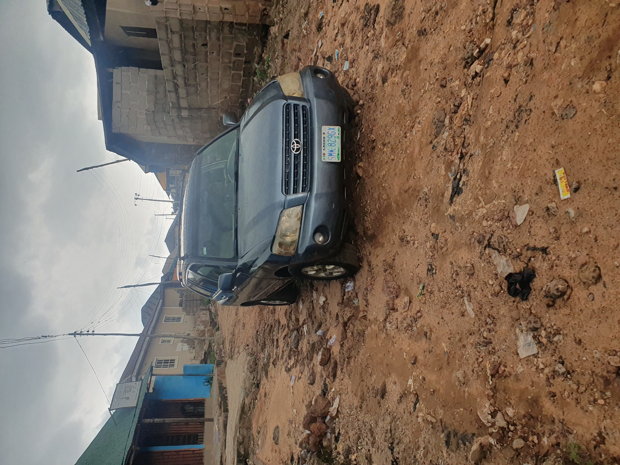 The image depicts a street scene in Lokoja, Nigeria, characterized by a heavily damaged and unpaved road surface filled with rocks, dirt, and debris. A dark blue Toyota Highlander SUV is parked on the right side of the frame, its front tire angled towards the camera. The vehicle's license plate, which reads "LAGOS SMK 829GX," is visible.
The surrounding environment consists of various buildings. On the left, a structure with a green roof and blue and brown walls is partially visible. To the right of the SUV, a building constructed from concrete blocks with exposed rebar and an open roof structure is present. The sky is overcast and cloudy, suggesting a potentially damp or rainy day. Electrical wires are strung across poles in the background, a common feature in urban Nigerian landscapes. The ground is littered with plastic bags, wrappers, and other refuse, indicating a lack of proper waste management. The overall impression is one of a neglected urban area with challenging road conditions.