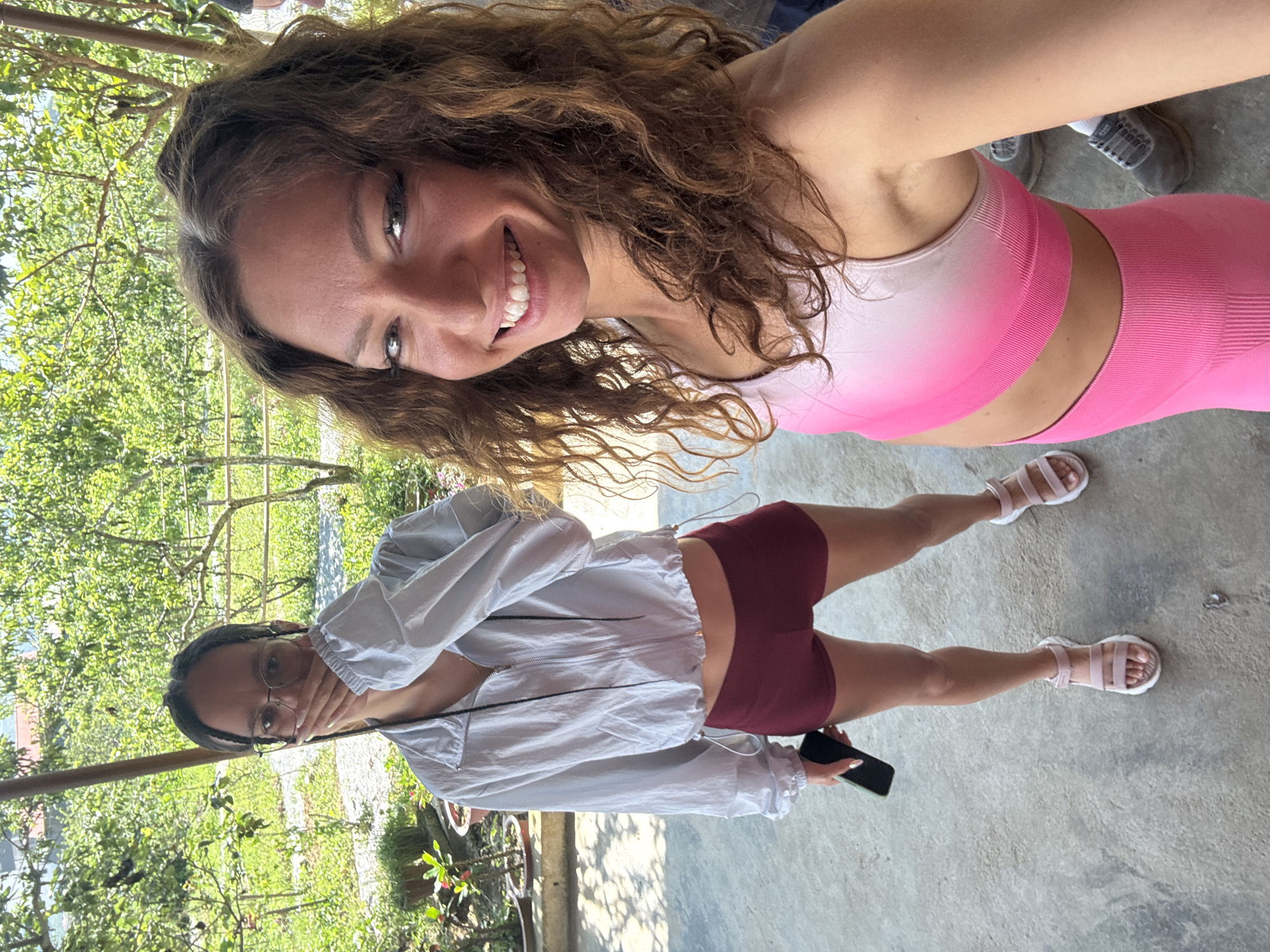 Two women are standing in an outdoor setting, possibly in Bắc Hà, Vietnam. One woman with curly brown hair is in the foreground, wearing a pink sports bra and leggings. She is smiling at the camera. Behind her, a second woman with dark hair and glasses is wearing a light-colored jacket and maroon shorts. She has one hand near her face and is holding a smartphone. The background shows greenery, including trees and potted plants, as well as elements of a building. The ground is paved. The image appears to be taken from a high angle.