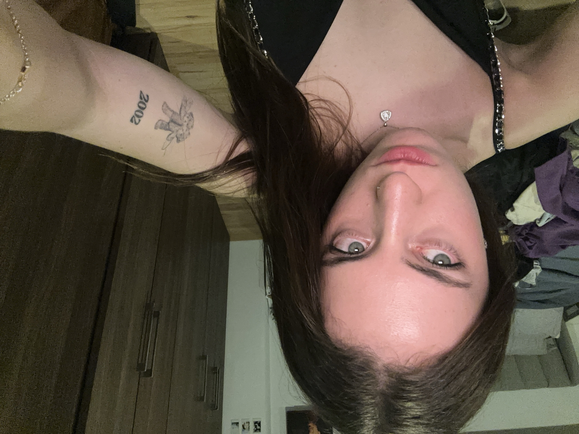 A young woman is posing for a selfie in an indoor setting. She has dark brown hair, light-colored eyes, and a nose piercing. She is wearing a black top and a delicate necklace with a heart-shaped pendant. Her left arm is visible and has a tattoo of "2002" and a small angel on the inner forearm. The background includes a dark wooden wardrobe, a white wall with small photographs, and some clothing items in disarray.
It is not possible to determine the location as Huixquilucan de Degollado, Mexico from the given visual information.