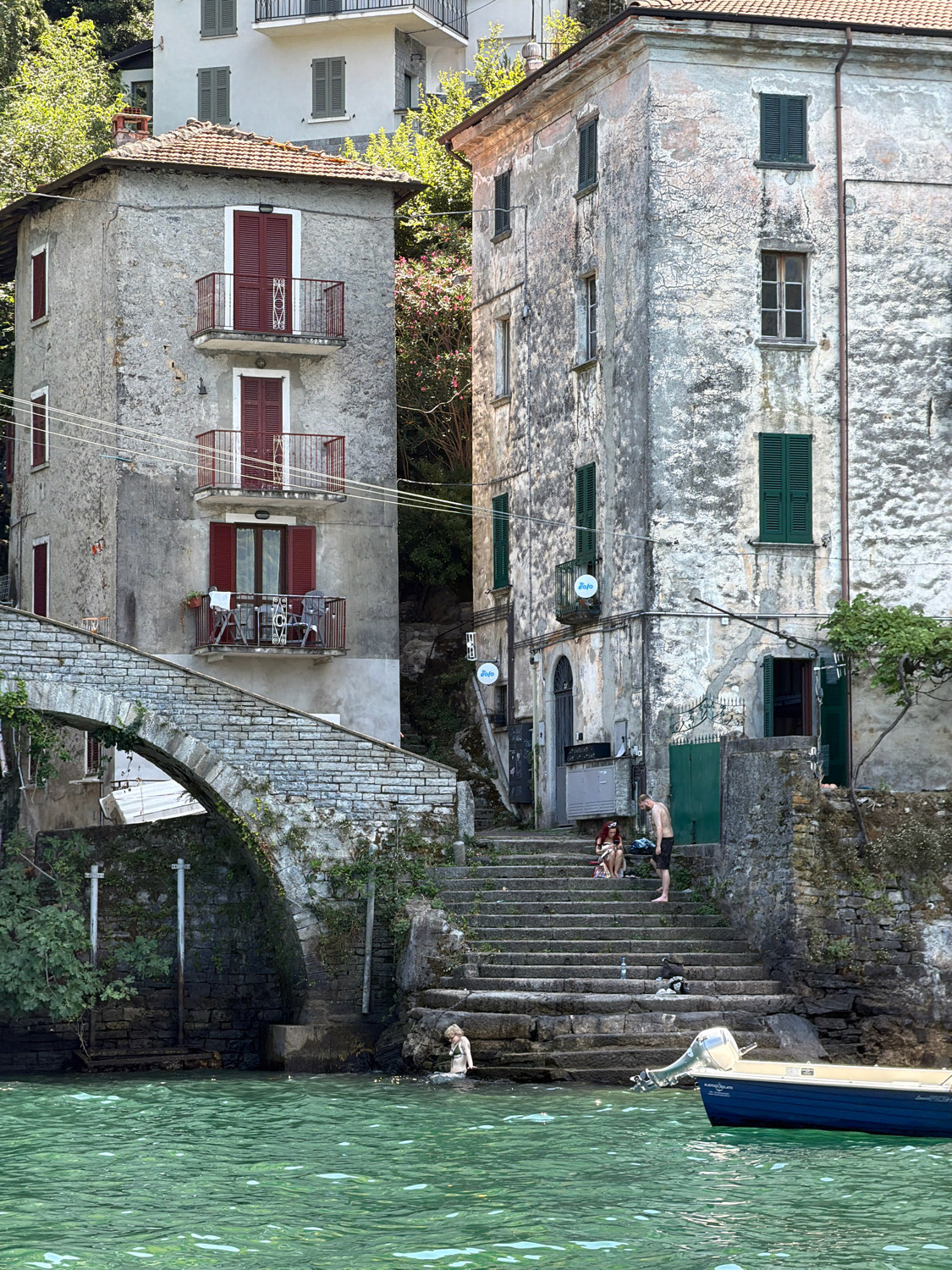 The image depicts a scene in Nesso, Italy, featuring old, weathered buildings situated along a body of water. On the left, a multi-story building with red shutters and balconies stands adjacent to a stone bridge arching over the water. Steps lead up from the water's edge to the buildings. In the foreground, the turquoise water is visible, with a small blue boat docked to the right. Several people are present: one person is partially submerged in the water near the steps on the left, and a group of three individuals are sitting and standing on the steps further up. The buildings themselves show signs of age, with peeling paint and textured facades. Lush green foliage surrounds the structures, indicating a natural, somewhat overgrown environment. The overall impression is of a historic lakeside village.