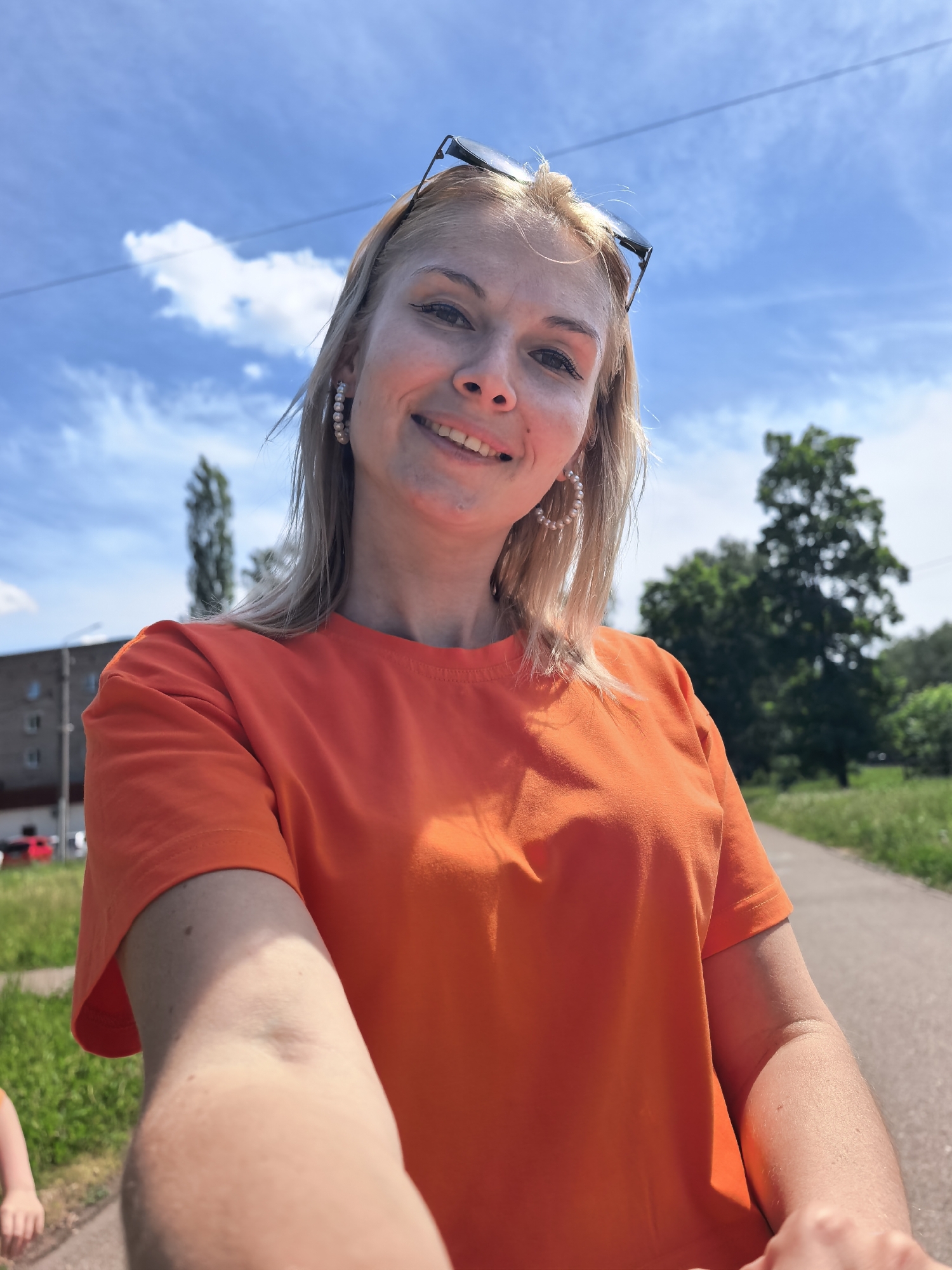A woman with blonde hair and wearing an orange t-shirt smiles into the camera, likely holding it at arm's length for a selfie. She has sunglasses perched on her head and pearl hoop earrings. The background shows a partly cloudy blue sky, a sidewalk, a tree, and a building. Based on the context provided (Sterlitamak, Russia), this is likely a public park or recreational area within the city.