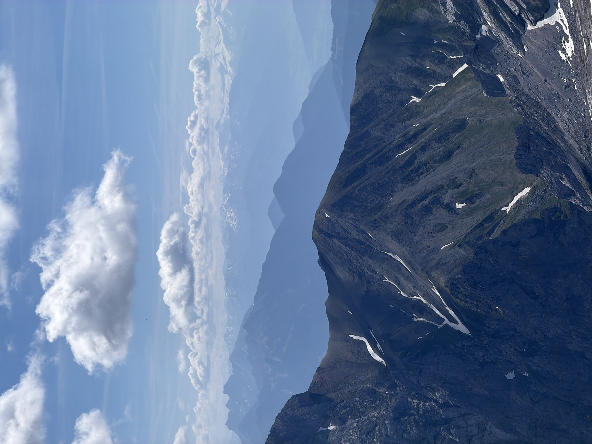 This is a vertical, high-angle, outdoor photograph of a majestic mountain range under a blue sky with fluffy white clouds. The foreground is dominated by a rugged, dark grey, rocky mountain peak. Patches of white snow are visible on its slopes, as are streaks of green vegetation.
In the mid-ground, a series of receding mountain ranges, rendered in progressively lighter shades of blue, stretch towards the horizon. The farthest ranges are misted and appear to have snow-capped peaks, suggesting a vast, expansive landscape. The transition of colors from dark grey in the foreground to pale blue in the distance creates a strong sense of depth.
The sky is a bright blue, with several large, cumulus clouds casting shadows on the mountains below. The overall lighting suggests it is daytime, likely midday or early afternoon.
While the image depicts a natural mountain landscape, it is not possible to determine the specific location within Garmisch-Partenkirchen, Germany, solely from the visual information provided. The scene is devoid of any human presence or man-made objects, focusing entirely on the natural grandeur of the mountains and sky.