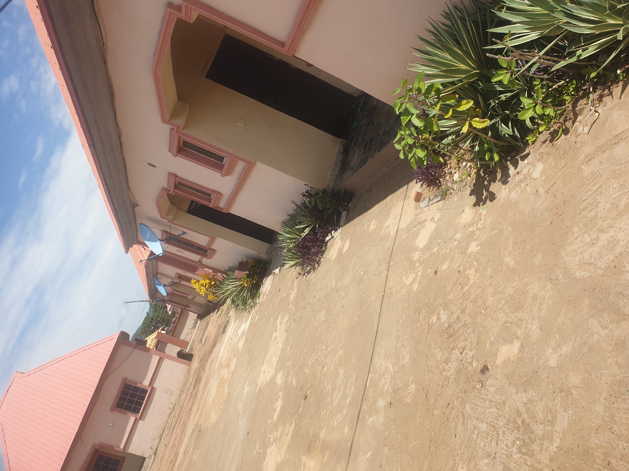 The scene depicts a row of single-story buildings in Lokoja, Nigeria. The buildings are painted in shades of beige and pink, with some having reddish roofs. Each building has windows, and some have doorways. The architectural style appears to be of standard residential design. Some structures are adorned with blue satellite dishes. Various plants and greenery grow alongside the buildings. The foreground is occupied by a paved area that looks like a driveway or walkway. The sky is blue with some wispy clouds. The lighting suggests it is daytime.