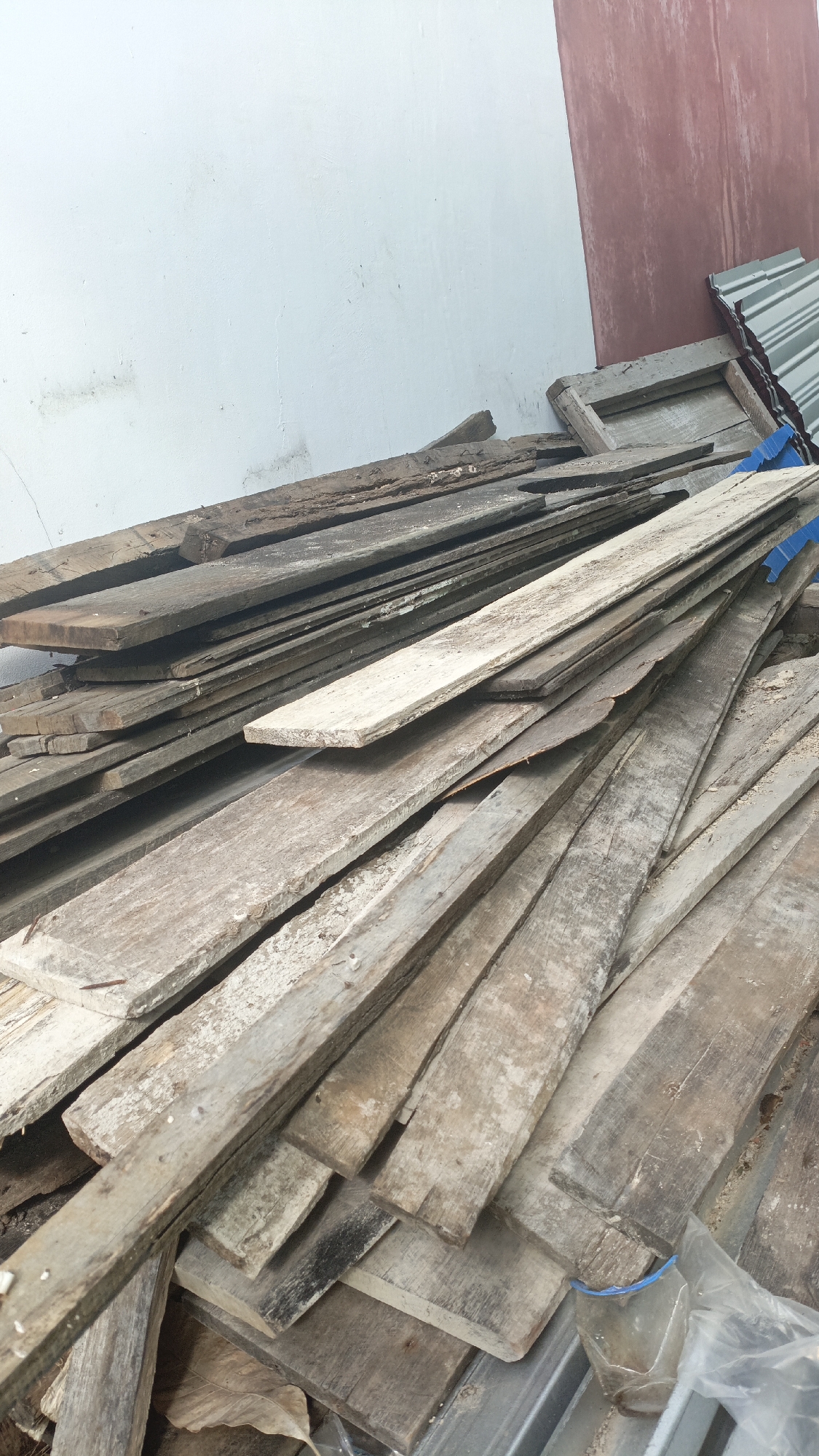 The image displays a pile of weathered, rough-hewn wooden planks. The planks are of varying lengths and widths, and they are stacked haphazardly. Some of the wood appears to be aged and has a gray, weathered patina, while other pieces are lighter in color. There are visible nail heads on some of the planks. In the background, a white wall is visible on the left, and a section of a reddish-brown wall is on the right. A corrugated metal roof is also visible at the top right of the image. The scene suggests an outdoor storage area, possibly at a construction site or a junkyard, in Saring Sungai Binjai, Indonesia.