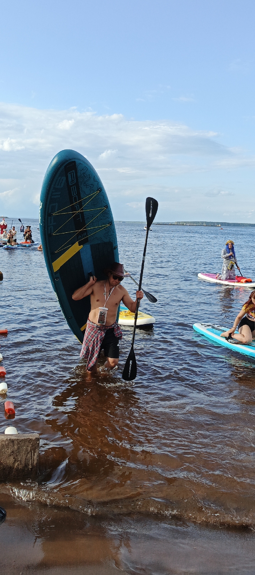 In Chaikovsky, Russia, on a sunny day with scattered clouds, a man is entering the water with a large, teal-colored paddleboard. He is shirtless, wearing black shorts, a red and black checkered shirt tied around his waist, sunglasses, and a brown hat. The paddleboard is propped upright on his right shoulder, with the paddle held in his right hand, angled towards the water. The man is partially submerged in the murky, reddish-brown water, with his legs visible below the surface.
Several other paddleboarders are visible in the background on the calm water. To the left, there are individuals dressed in what appear to be costumes, including a Santa Claus. To the right, two individuals are on paddleboards; one is kneeling and wearing a blue head covering and striped shirt, while another is also kneeling and wearing a colorful top. The water extends to a distant horizon with land and buildings visible. The scene appears to be at a waterfront area with a concrete edge in the foreground on the left. Several red and white buoys are floating near the shore.