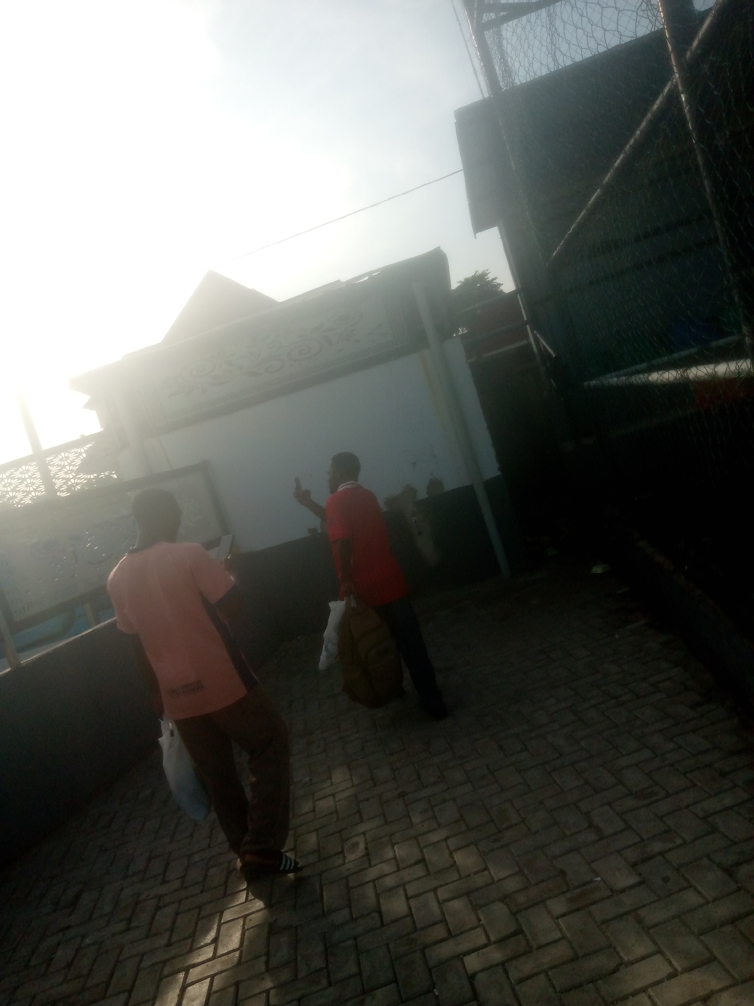 In Jalingo, Nigeria, two individuals are captured in a street scene. One person, wearing a pink t-shirt and brown trousers, is walking away from the camera, holding a white plastic bag. The second person, in a red t-shirt and dark trousers, is facing away from the viewer and appears to be holding a mobile phone. They are standing on a paved walkway, possibly in a public area or near a building. The background shows a wall with decorative elements and a fence, suggesting an outdoor environment. Sunlight is a prominent feature, casting shadows on the ground and creating a bright, slightly overexposed atmosphere.