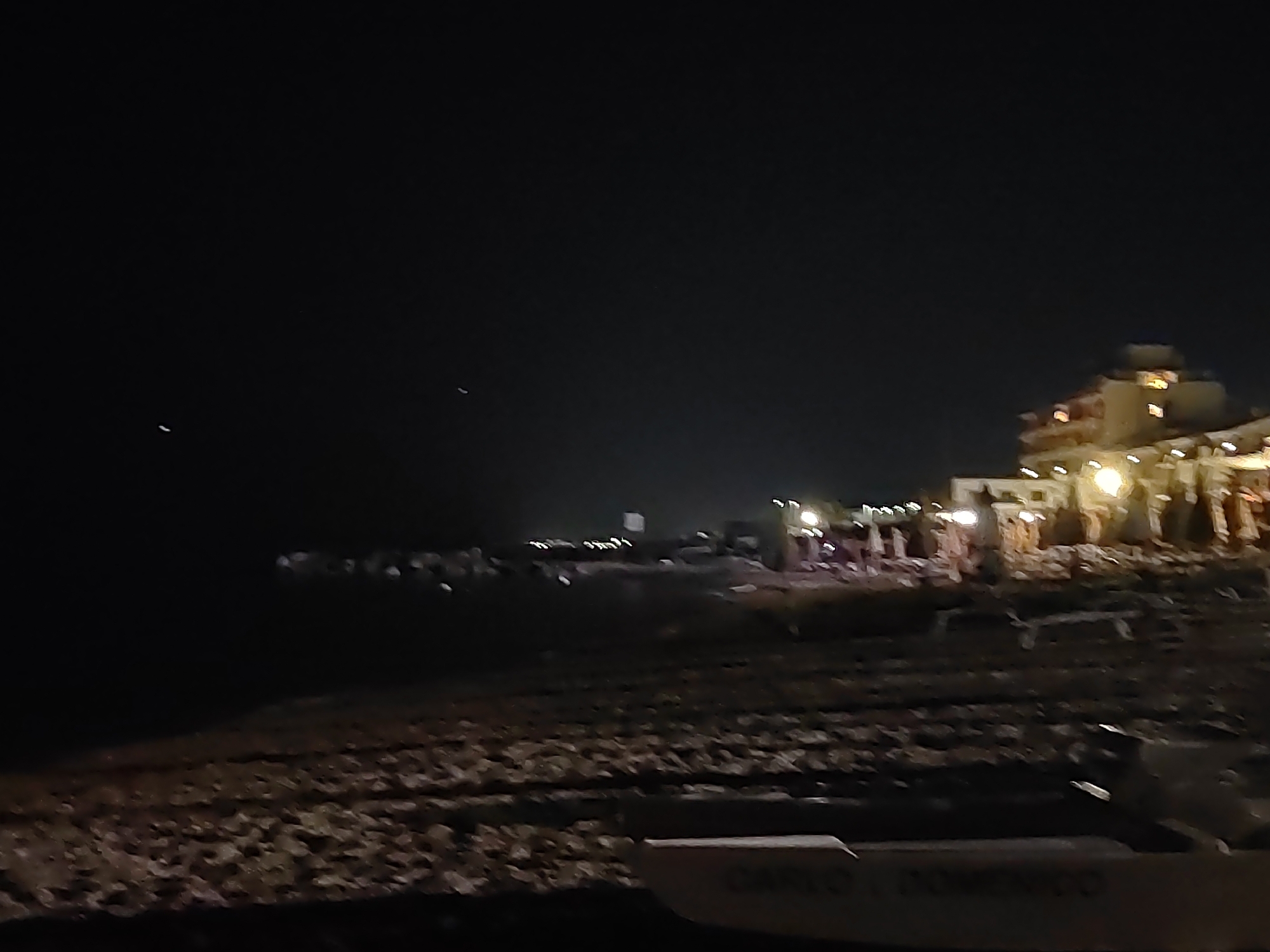 The image captures a dimly lit nighttime scene at the beach in Porto Recanati, Italy. A body of water, likely the Adriatic Sea, is visible on the left, with faint reflections of lights on its surface. The shore in the foreground is covered in pebbles and debris. In the background, illuminated structures suggest a beachfront area with buildings and lights, possibly restaurants or hotels. A white object, possibly a boat or debris, is partially visible in the lower right corner, with some faint lettering on it. The overall atmosphere is dark and somewhat grainy, indicating low light conditions and possibly a long exposure. There are no identifiable people or specific actions depicted.