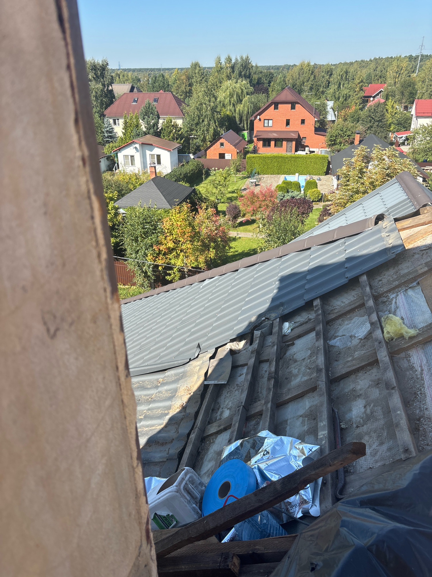 An overhead shot captures a section of a damaged roof in Zhavoronki, Russia, with the surrounding residential area visible in the background. The foreground is dominated by the exposed wooden rafters and insulation of the roof, with some construction materials like rolls of blue tape, a white plastic container, and a silver foil-wrapped object scattered on top. The damaged roof area appears to be under repair or renovation. Beyond the immediate roof structure, several houses with red and white facades and dark roofs are nestled among lush green trees. The sky is clear and blue, indicating fair weather.