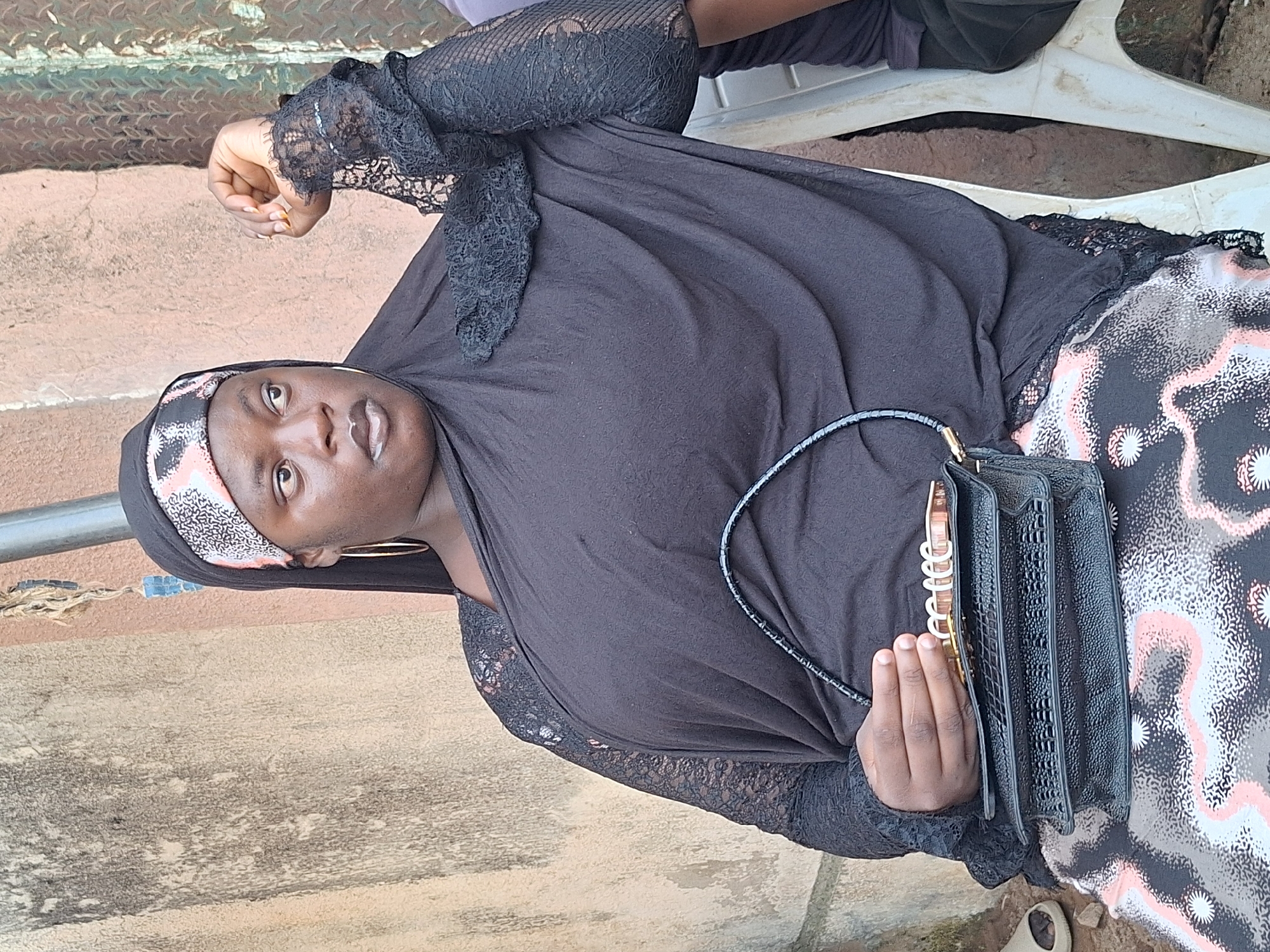 A medium shot captures a Black woman sitting on a white plastic chair in Narayi, Nigeria. She is wearing a black hijab with a patterned band, a black abaya with black lace sleeves, and a patterned skirt. She holds a black clutch purse in her lap with her left hand, and her right arm is bent with her hand resting on her arm. She is looking to her left with a neutral expression. The background consists of a textured wall and a corrugated metal surface.