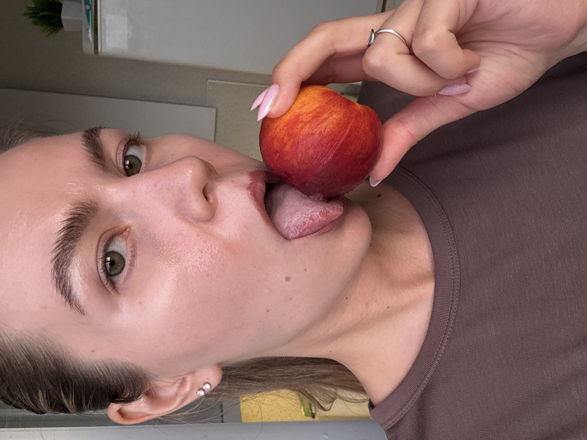 An extreme close-up photograph shows the face of a young woman with light skin and dark eyebrows, holding a peach up to her mouth with her right hand. Her tongue is extended and touching the peach. She is wearing a dark brown t-shirt. The background is out of focus, showing a white appliance and a small potted plant. The lighting is soft and natural. There are no identifiable city or location details.