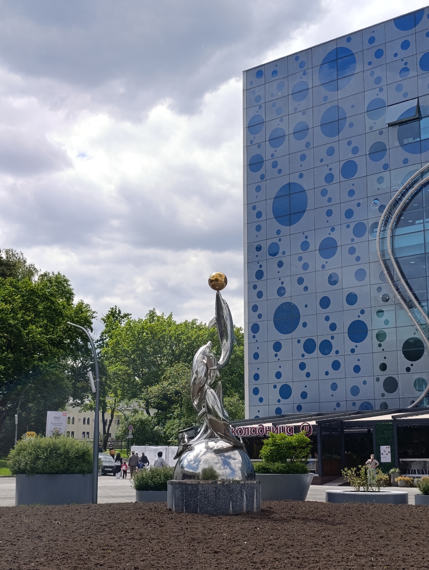 This outdoor photograph depicts a modern urban scene in Moscow, Russia, under an overcast, cloudy sky. In the foreground, a prominent, highly reflective metallic sculpture captures attention. It features two abstract, flowing forms, possibly aquatic creatures like fish or dolphins, supporting a golden sphere at their apex. The sculpture rests on a dark, polished stone base, situated in a patch of dark, freshly tilled earth.
Behind the sculpture and to the right stands a large, multi-story building with a distinctive light-colored facade. This building is adorned with numerous blue circles of varying sizes, resembling bubbles, which are scattered across its surface. It features large windows, some of which are reflective, and a unique, curved architectural element made of a mesh-like material on its right side. Below the building's facade, an outdoor seating area with tables is visible, and the red Cyrillic text "коладница" (Koladnitsa), likely indicating a cafe or restaurant, is prominently displayed above an entrance.
To the left, lush green trees with dense foliage form a natural backdrop. A street or pedestrian pathway runs through the mid-ground, where a lamppost stands and a dark car is partially visible. Several indistinct figures of people are seen walking along this path and near the building's entrance. The scene suggests a public space, possibly a plaza or area surrounding a modern attraction like an aquarium, given the building's bubble motif. The time of day is daytime, with soft, diffused light from the cloudy sky.
