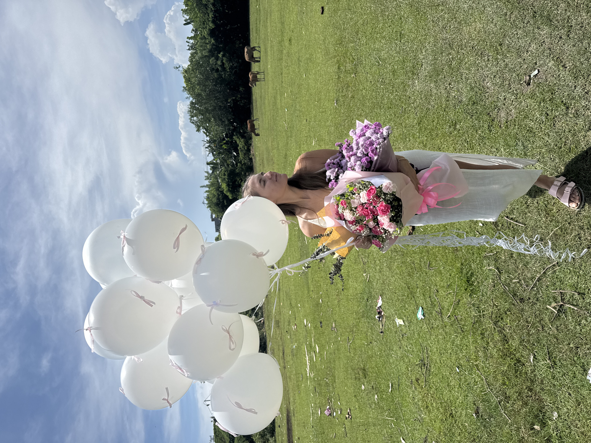 A young woman is lying on a grassy field, holding two bouquets of flowers and a bunch of white balloons. She is wearing a light-colored dress and sandals. The field is green and there are some cows grazing in the background, near a line of trees. The sky is blue with some white clouds. The location is likely in a rural area of Unknown, Unknown.