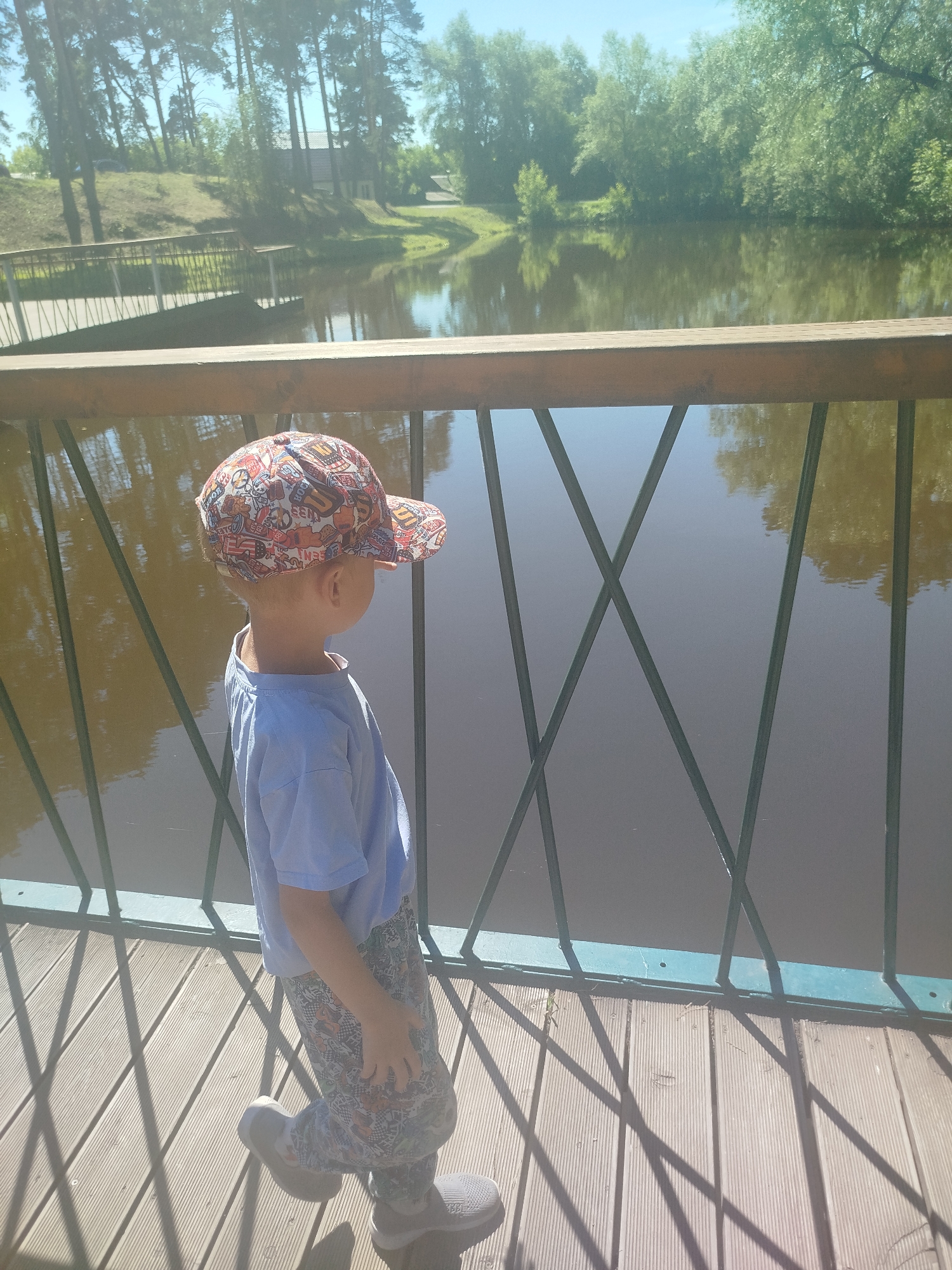 In Zavodoukovsk, Russia, a young boy stands on a wooden bridge overlooking a tranquil body of water. The boy is wearing a colorful, patterned baseball cap, a light blue t-shirt, and patterned leggings. He is positioned in the foreground, facing away from the camera and towards the water. The bridge has a wooden plank surface and a railing with green metal supports that create a crisscross pattern. The water reflects the surrounding trees and the clear blue sky. Beyond the water, lush green trees and vegetation line the far bank, with a hint of buildings and a pathway visible in the distance. The sun casts long shadows from the bridge railing onto the wooden planks.