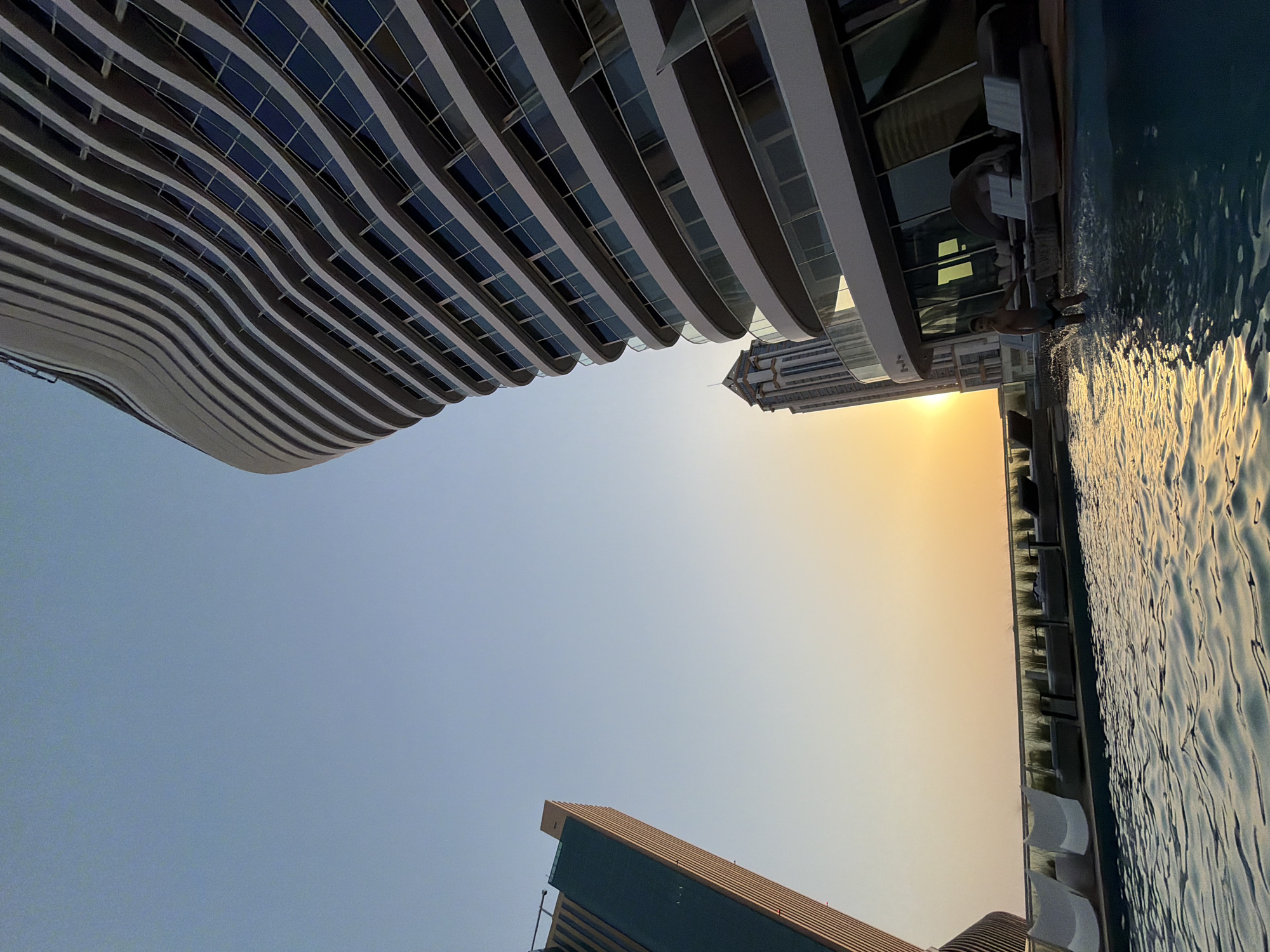 In the city of Lusail, Qatar, a rooftop swimming pool reflects the setting sun. The water is disturbed by a young male wading at the edge of the pool, his body angled towards the right side of the frame. He is wearing dark swim trunks. In the background, a modern, undulating skyscraper with wavy balconies dominates the right side of the image. To the left, another tall building with a green facade and a reddish-brown top is visible. Further in the distance, a more traditional skyscraper with a tiered top stands against the orange and blue gradient of the sunset sky. The pool area features several lounge chairs, some white and some dark gray, arranged along the edge of the pool, behind a glass barrier. The lighting suggests it is late afternoon or early evening, with the sun casting a warm glow on the scene.