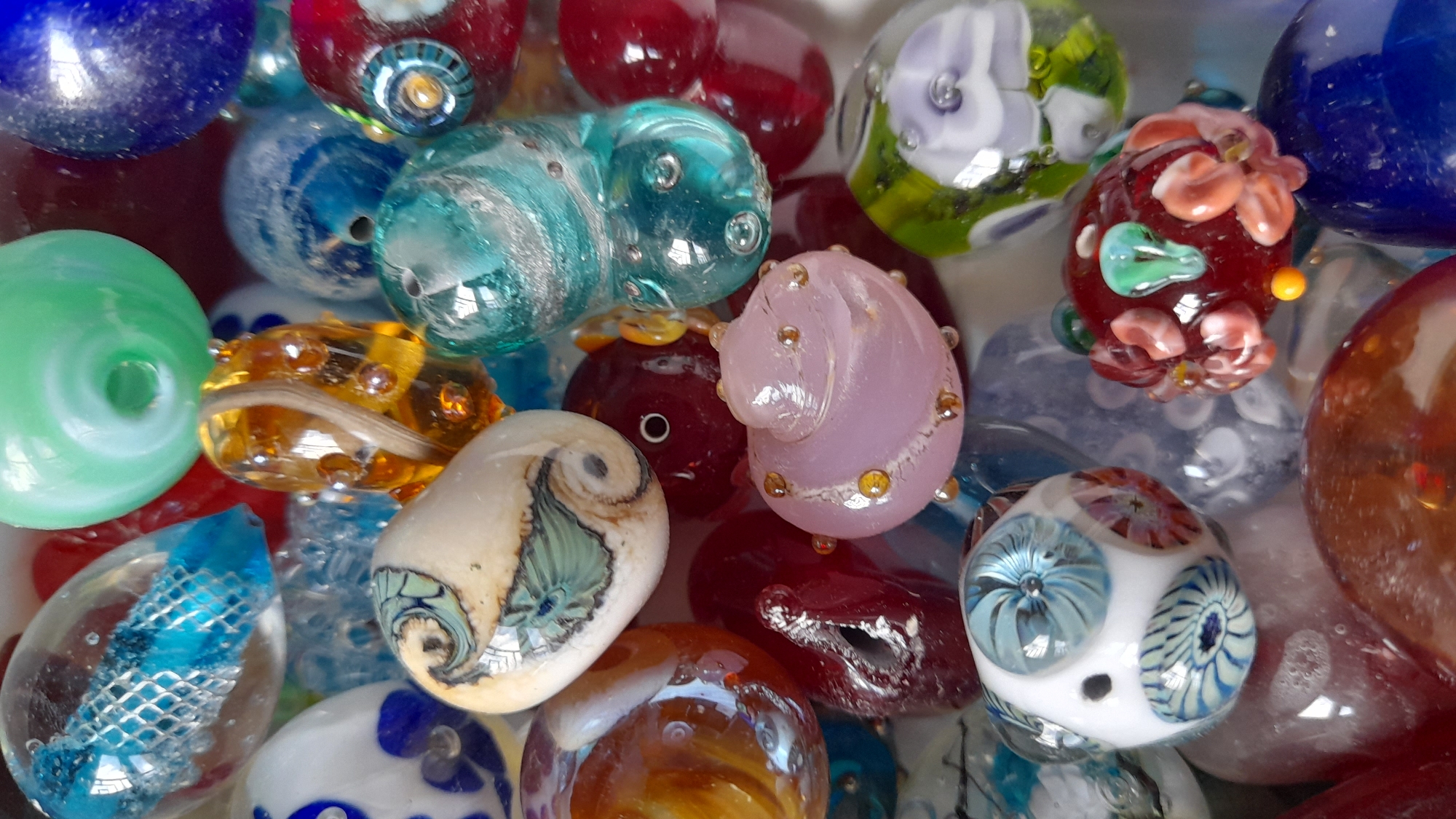 The image shows a close-up of a collection of glass beads. The beads are of various colors, shapes, and designs. Some are round, others are oval, and some have unique patterns. The colors include blue, red, green, yellow, pink, white, and brown. Some of the beads are clear with internal designs. The overall impression is one of a colorful and diverse assortment of handcrafted glass beads. The location is not provided in the image.